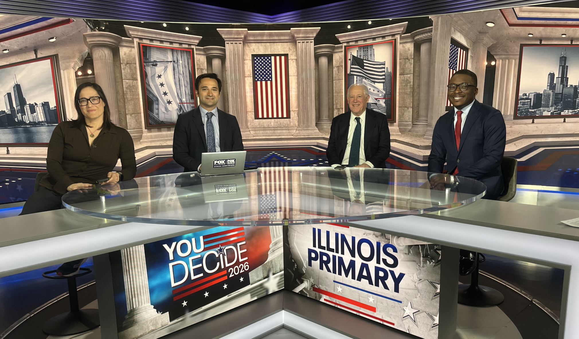 Four news anchors sit behind a curved glass desk in a patriotic studio with flags and columns; on-screen banners read "YOU DECIDE 2026" and "ILLINOIS PRIMARY".