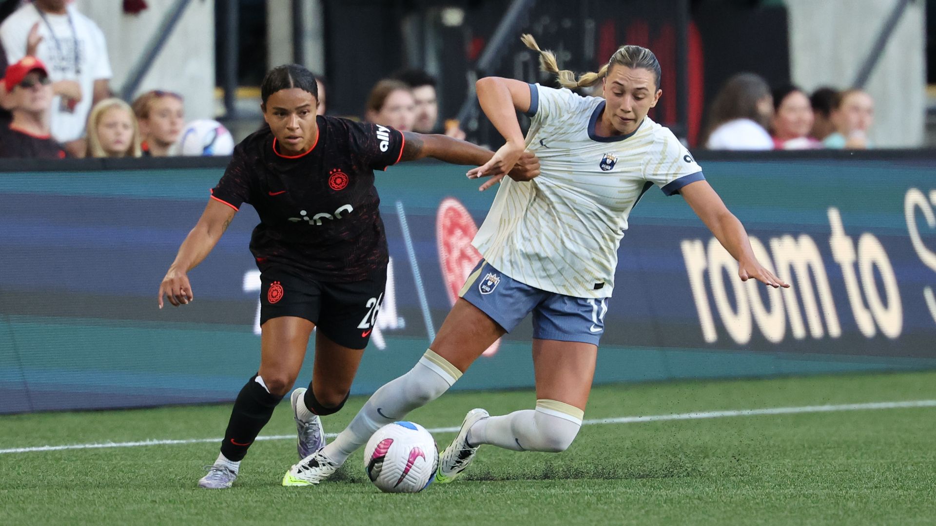 Two female soccer players contesting the ball on green field; one in black jersey with red trim, the other in white and blue jersey, with blurred spectators in background.