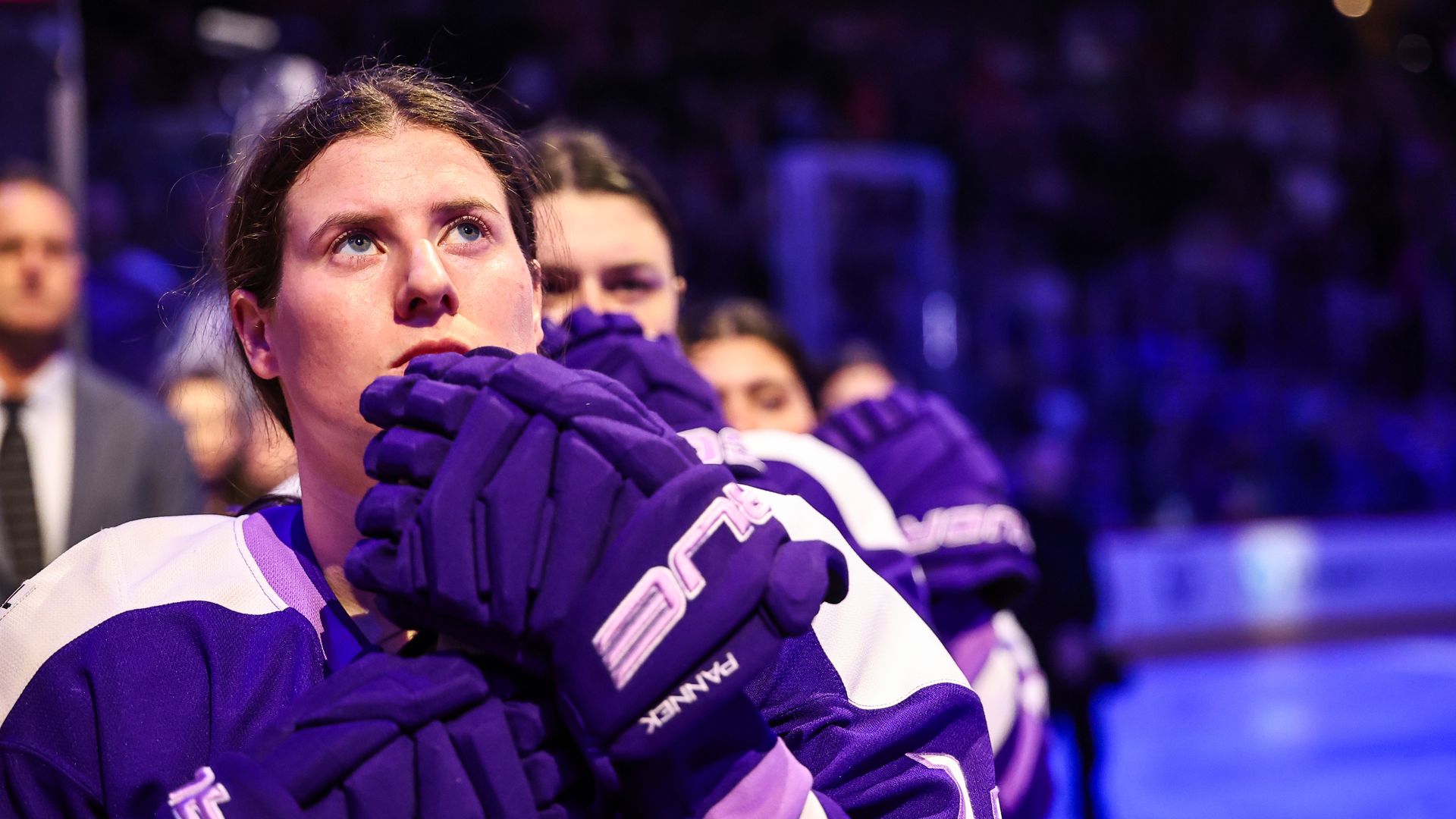 Close-up of a hockey player in a white and purple sports jersey wearing thick purple gloves, looking upward as a blurred crowd watches in a dim arena with neon blue and red lights.