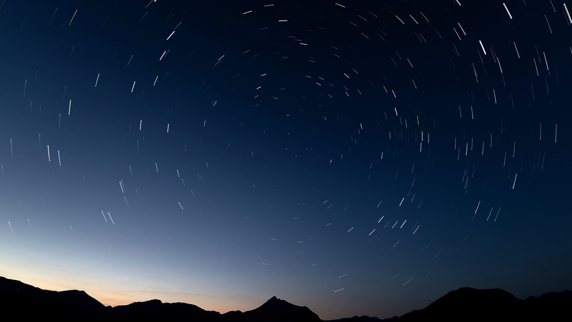A long-exposure photograph shows  circular star trails across a darkening sky above a silhouette of mountain peaks. 