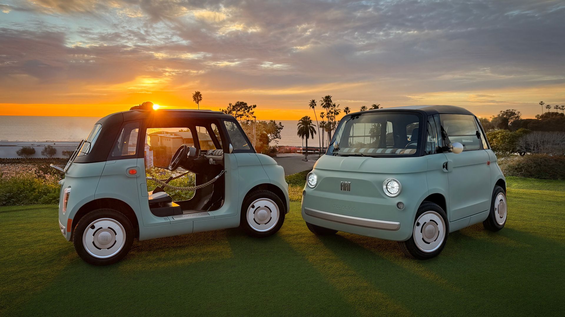 Two tiny, light blue Fiat Topolino electric cars parked on a coastal golf course at sunset, with the sun low on the horizon, orange sky, palm trees in the distance. The car on the left has the doors removed.