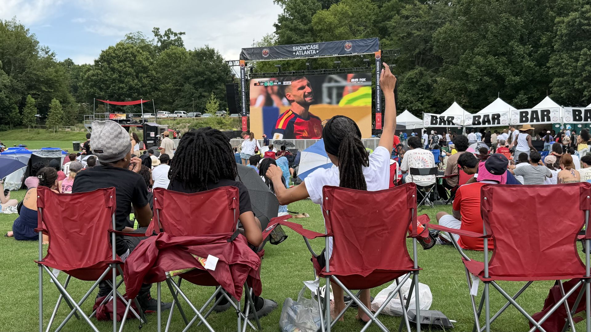 Outdoor event with a large screen showing a soccer match. Crowd sits in red folding chairs on grass; one person raises a hand. White BAR tents line the right; a banner reads Showcase Atlanta.