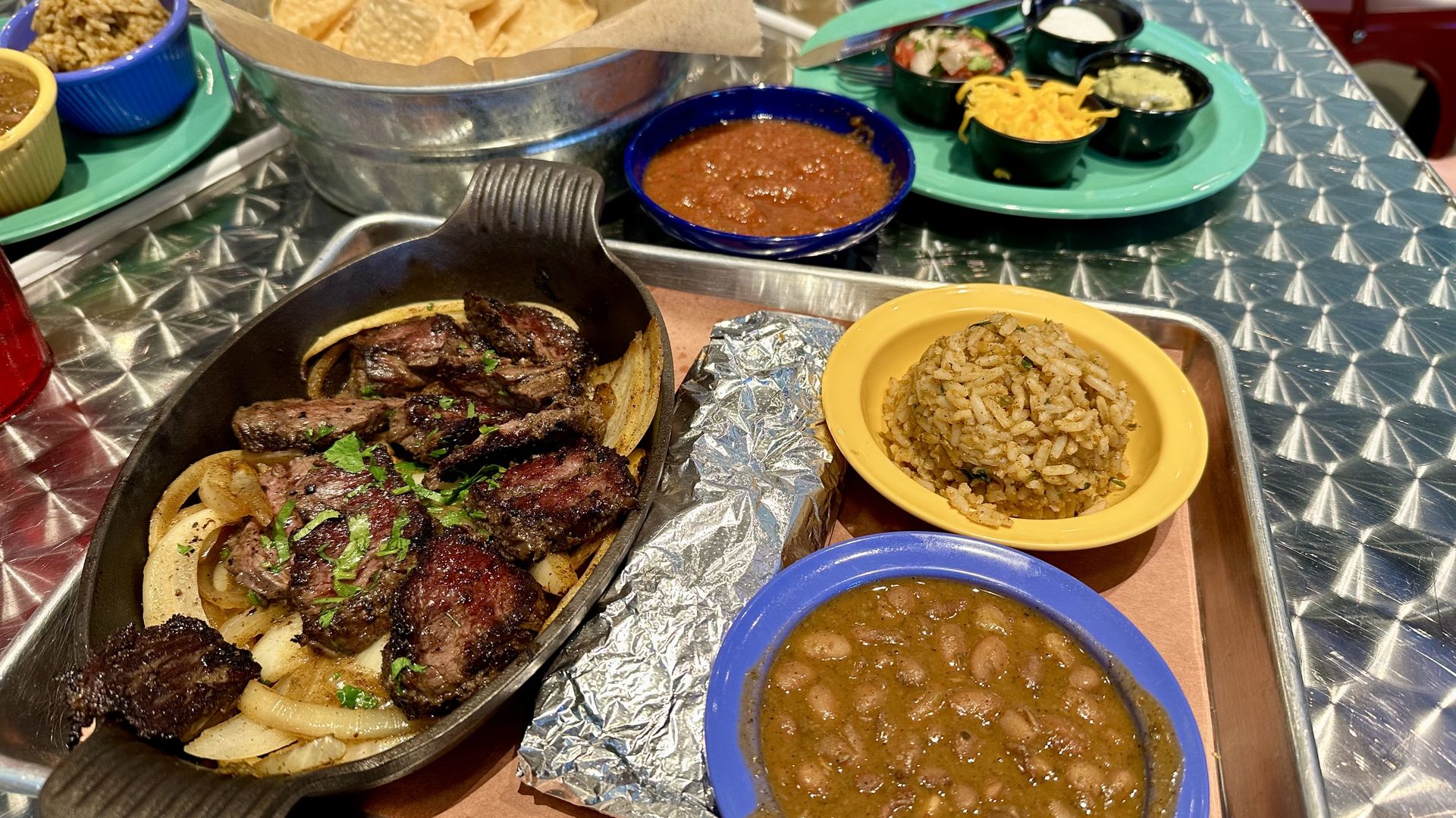 Sizzling steak fajitas with onions and cilantro in a cast-iron skillet on a metal table, with beans, salsa, yellow rice, chips, and toppings on a green platter.