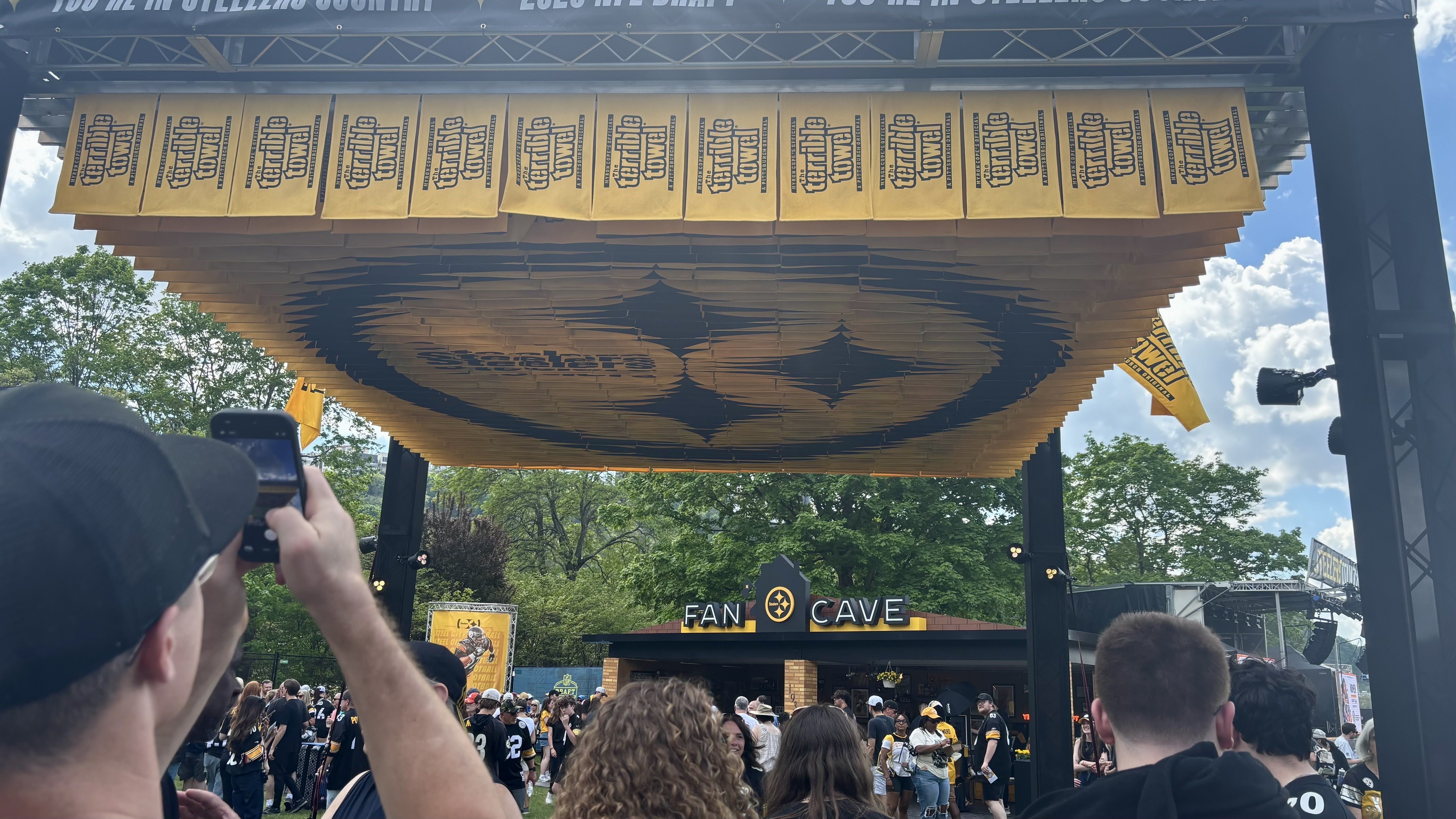 Outdoor festival scene with a massive gold and black layered canopy and banners above a crowd in black and gold. A building reads "FAN CAVE" in the background on a sunny day.
