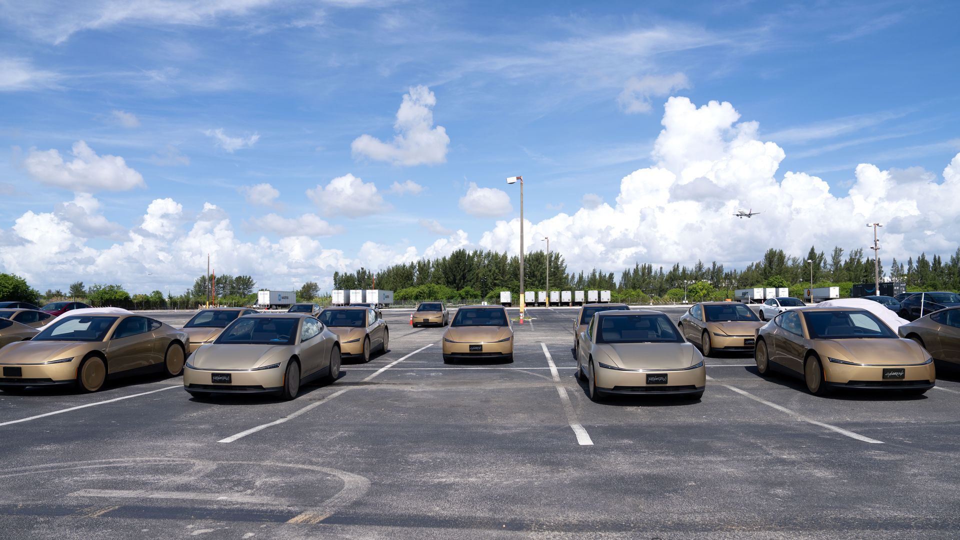 A parking lot full of gold-colored Tesla Cybercabs in neat rows under a clear blue sky with fluffy white clouds; distant trees line the horizon, with a small airplane visible in the distance.