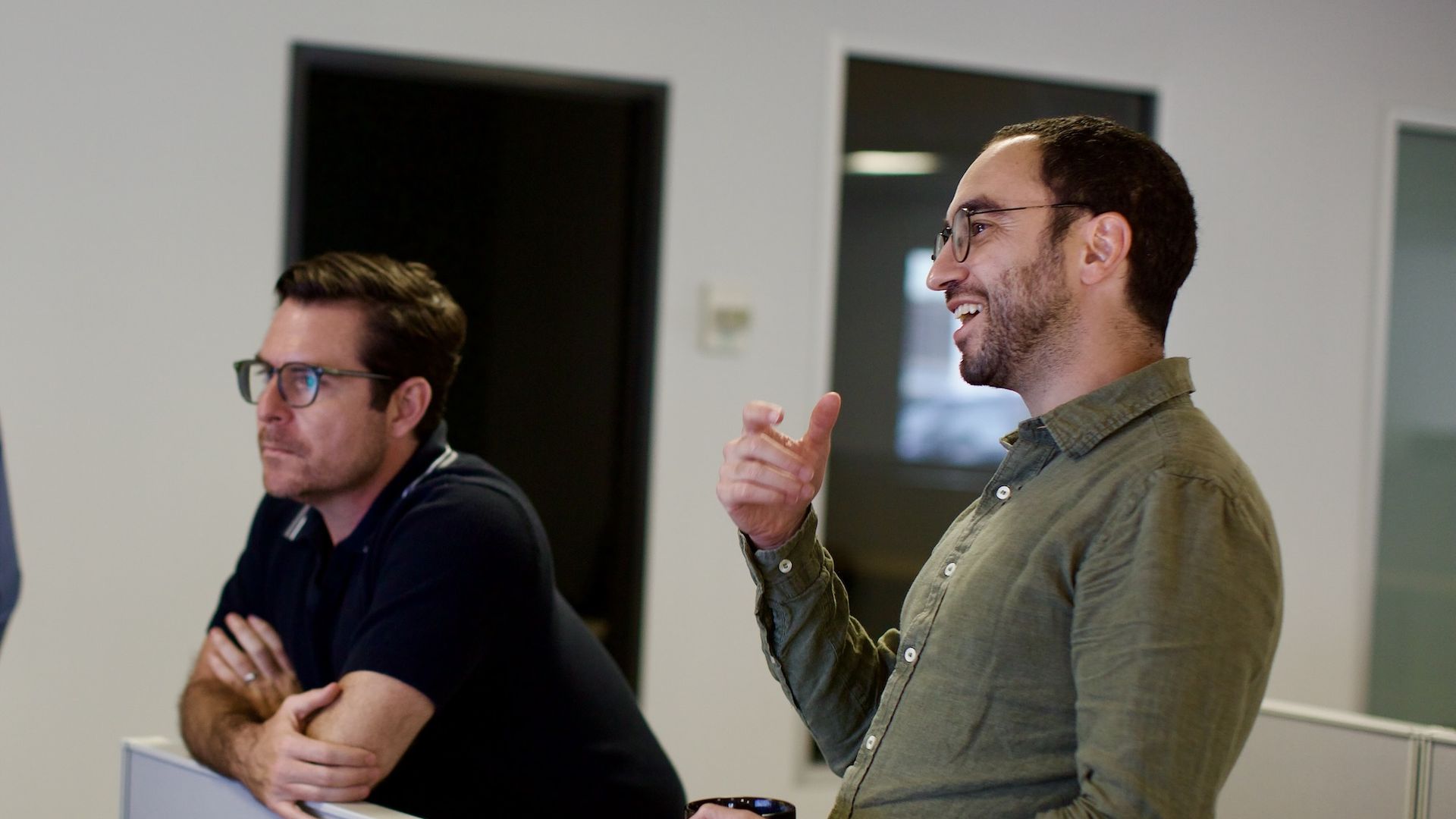 Two men with glasses having a conversation in an office, one wearing a dark polo shirt leaning on a cubicle wall, the other in a green button-up shirt holding a mug and gesturing.