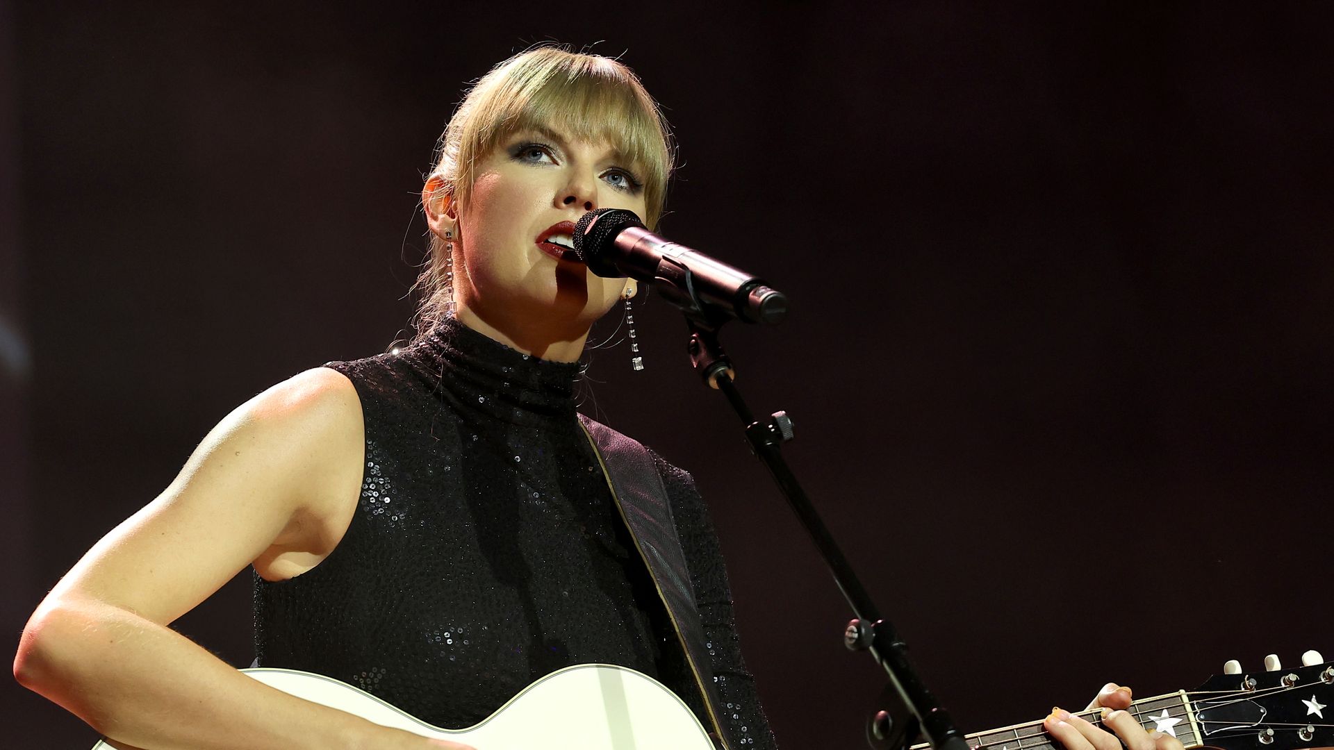 Taylor Swift plays guitar while singing into a microphone.