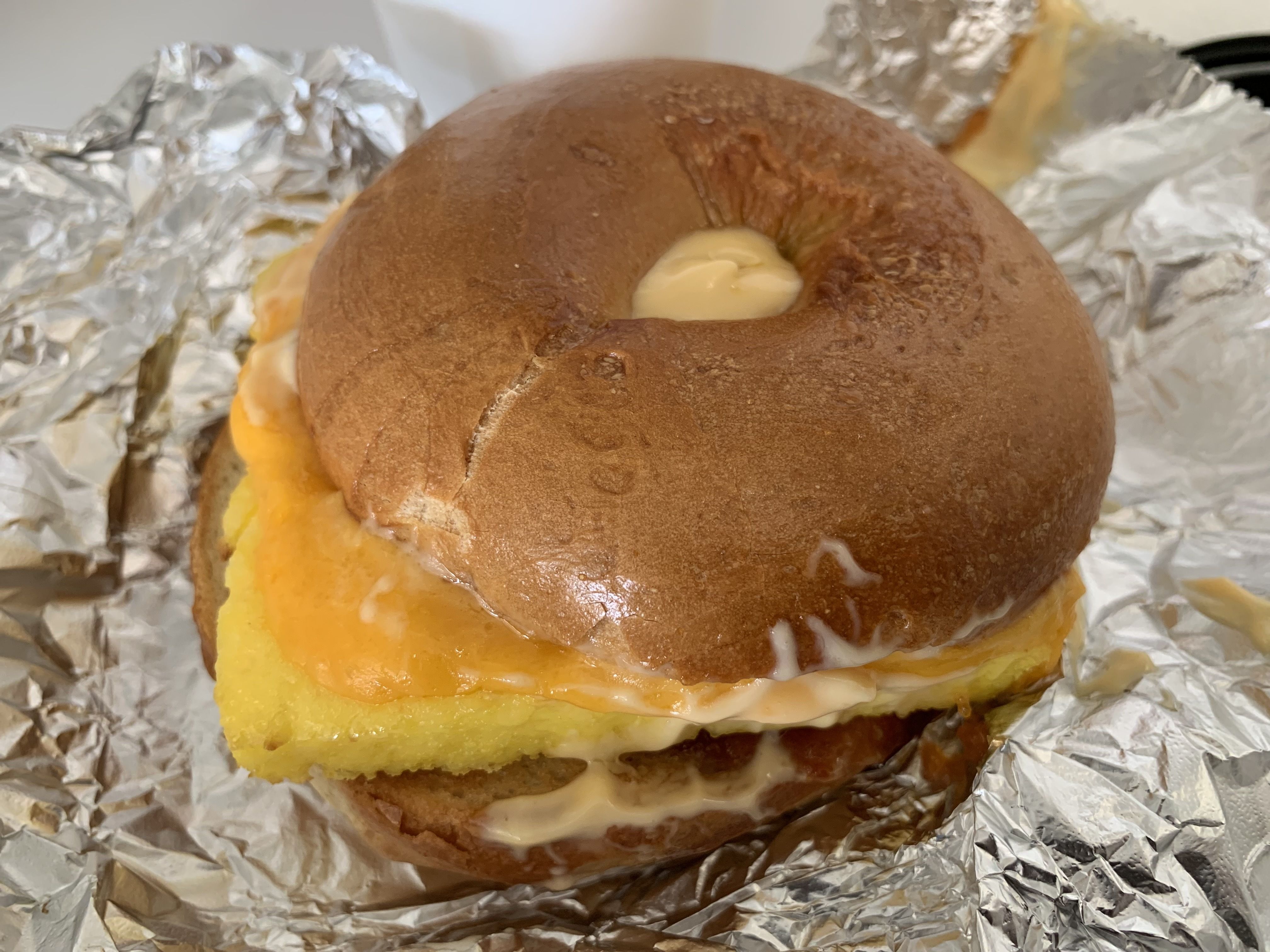 Macro photo of a round brown bun sandwich with melted yellow cheese and white sauce seeping out, surrounded by crumpled aluminum foil.