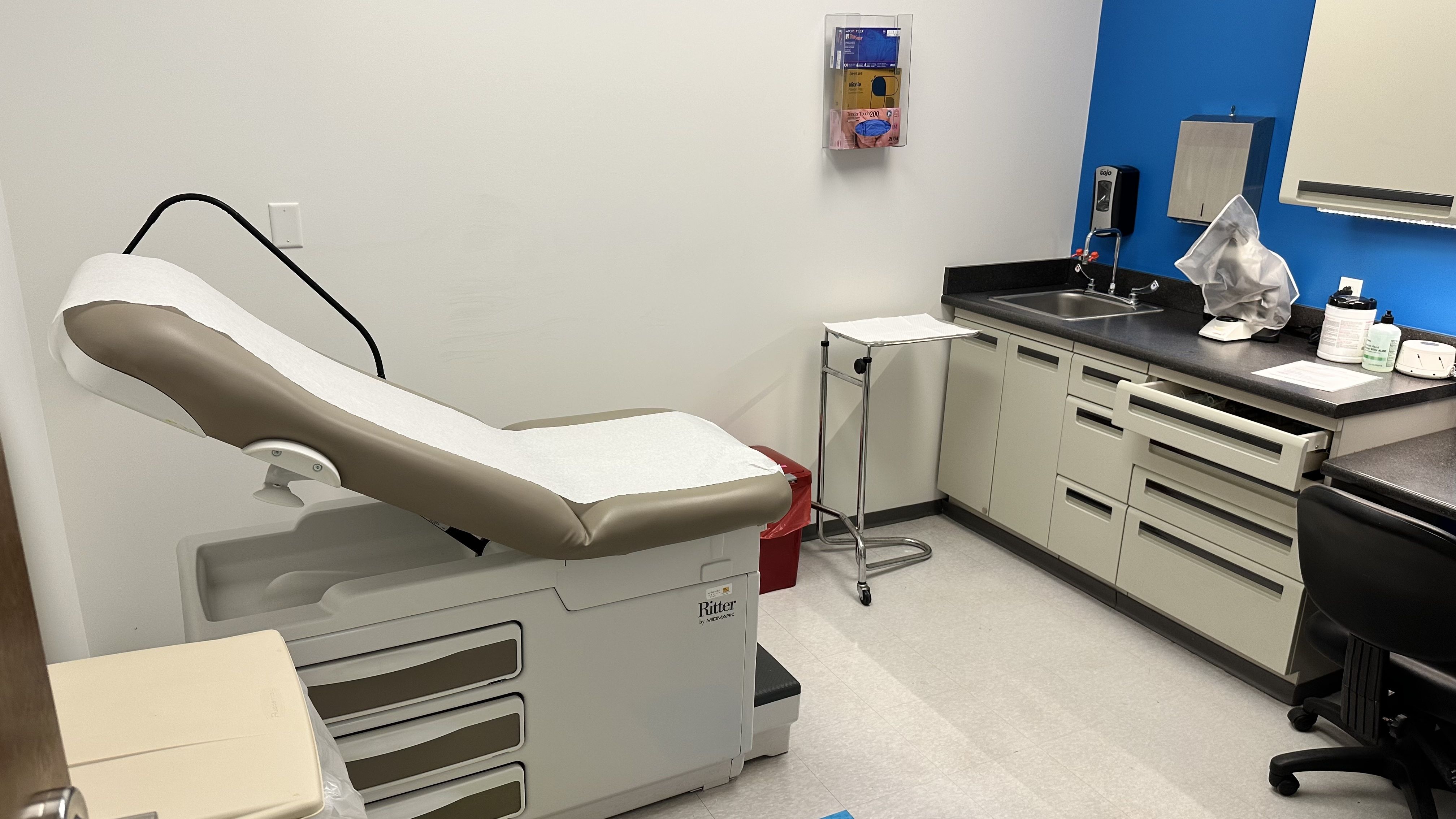 Medical exam room with a beige reclining exam chair covered in white paper, a chrome stand, white and gray cabinetry, a blue accent wall, a sink with faucet, and assorted supplies on the counter, including a covered device.