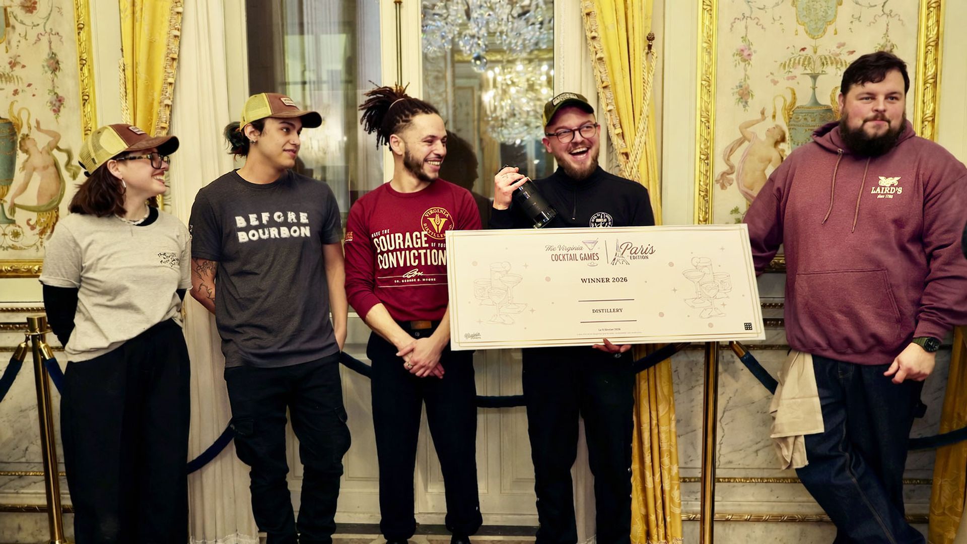 Five people in casual attire stand in a decorated room with gold curtains. One holds a large sign reading "The Virginia Cocktail Games Paris Edition WINNER 2026 DISTILLERY."