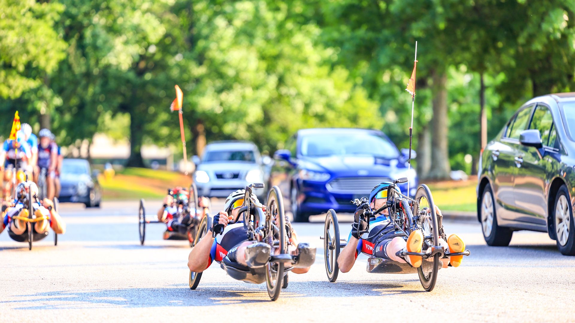 Several para-cyclers ride recumbent bicycles down a street as cars pass by them in a wooded setting.