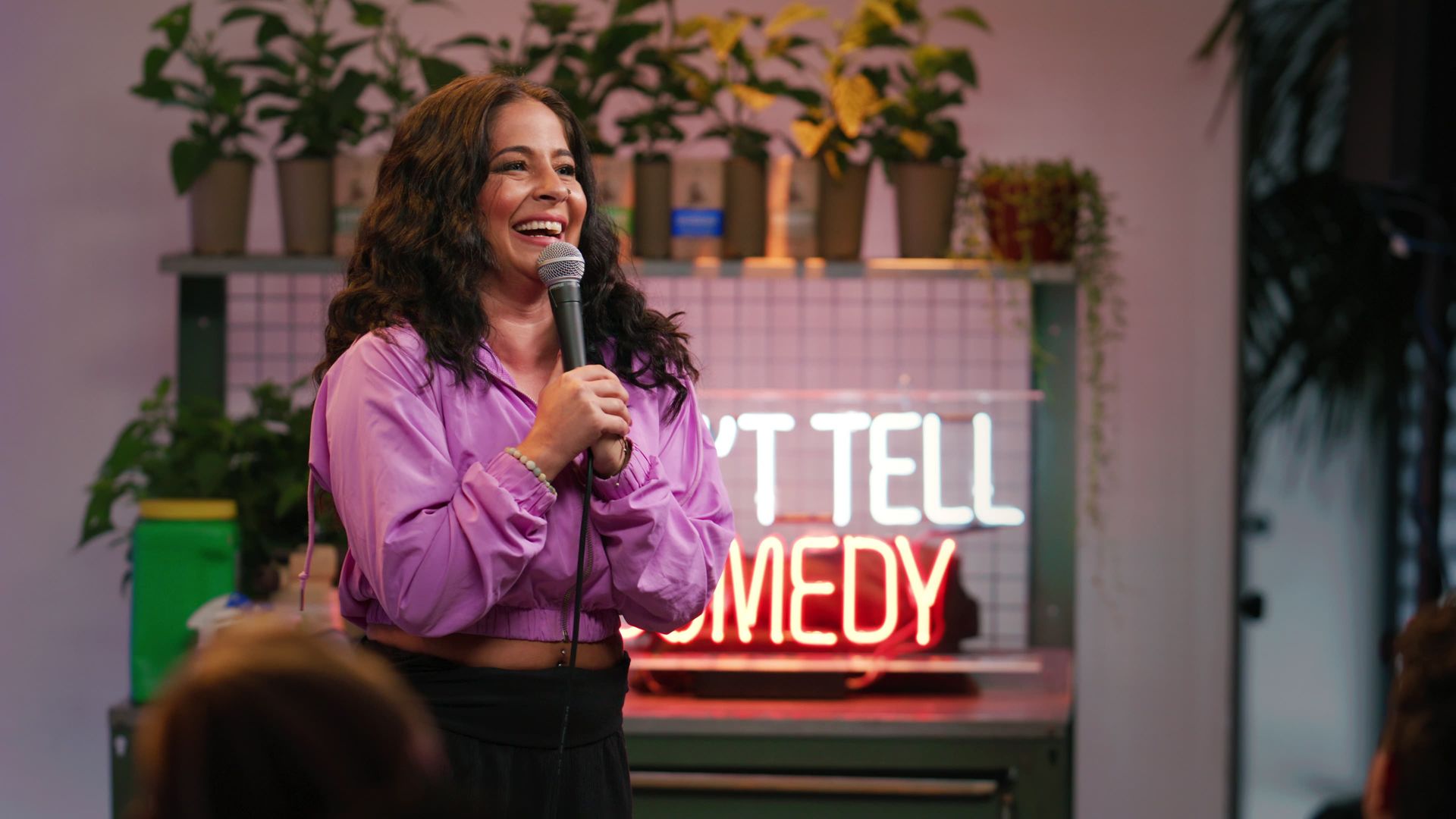 Smiling woman on stage holding a microphone, wearing a light purple jacket. Background shows potted plants and a neon red sign that reads "COMEDY", suggesting a casual comedy club setting.