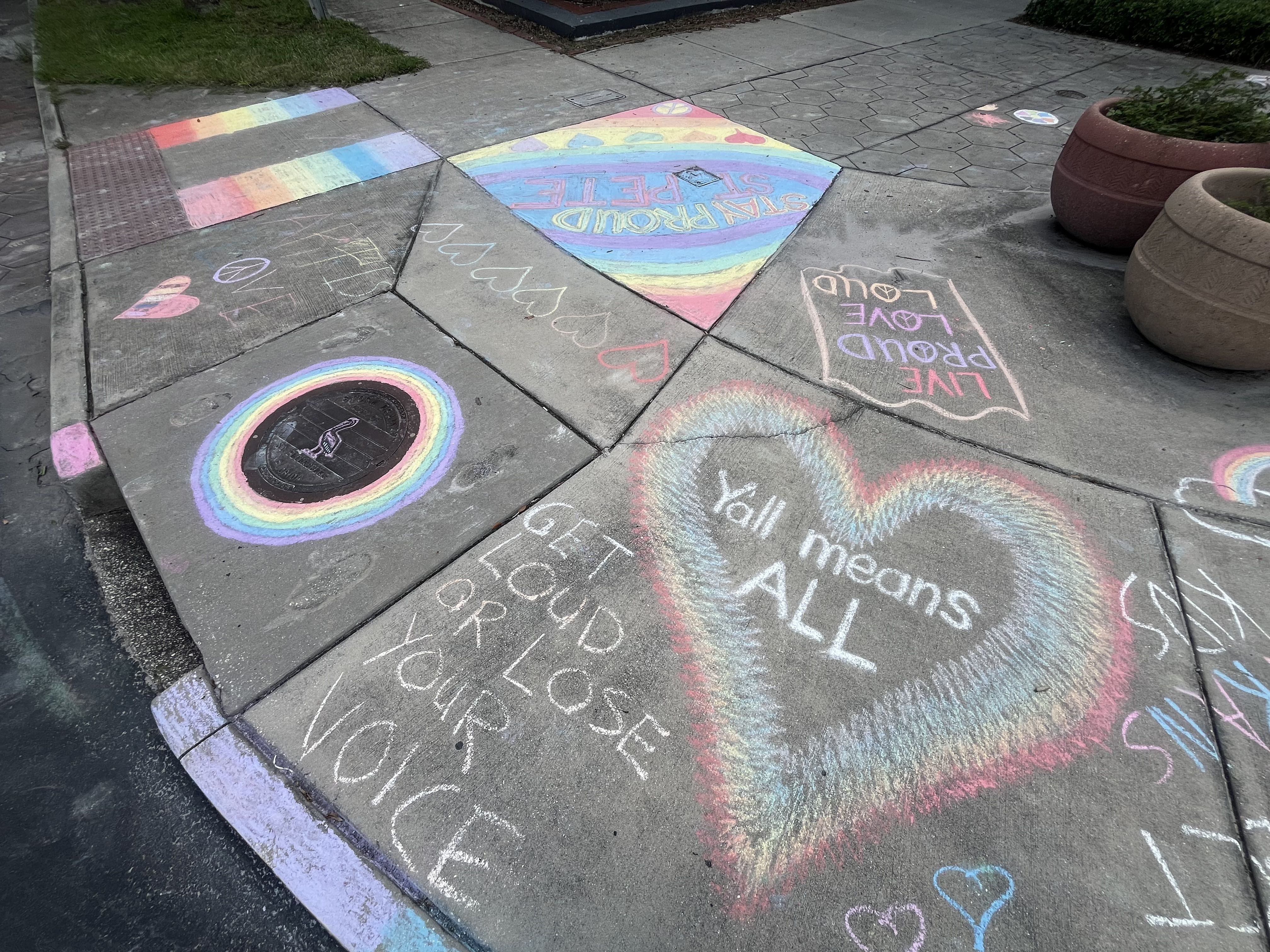 Sidewalk chalk art with rainbow flags, hearts, and messages like "Y'all means ALL" inside a rainbow heart and "GET LOUD OR LOSE YOUR VOICE" promoting pride and inclusivity.