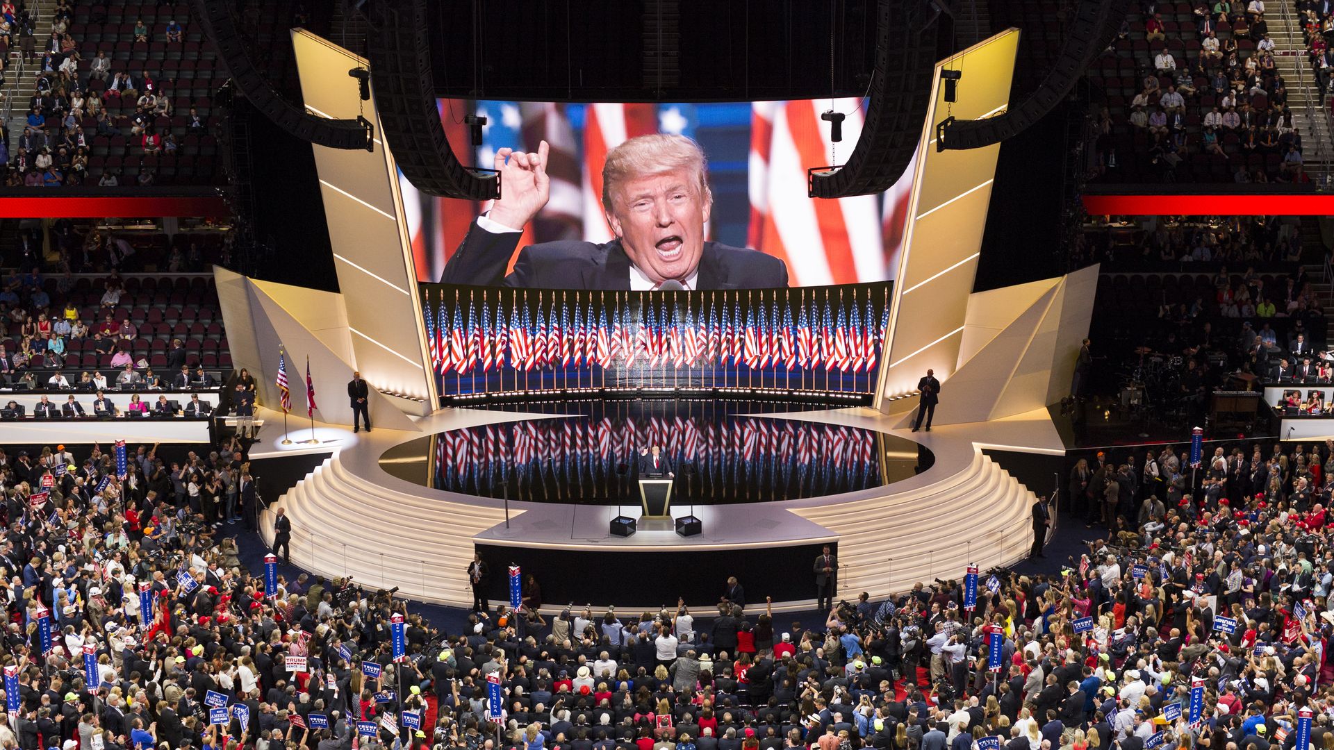Large crowd with American flags and signs at a political convention watching a speaker on stage with Donald Trump's image on a large screen behind, patriotic red, white, and blue colors.