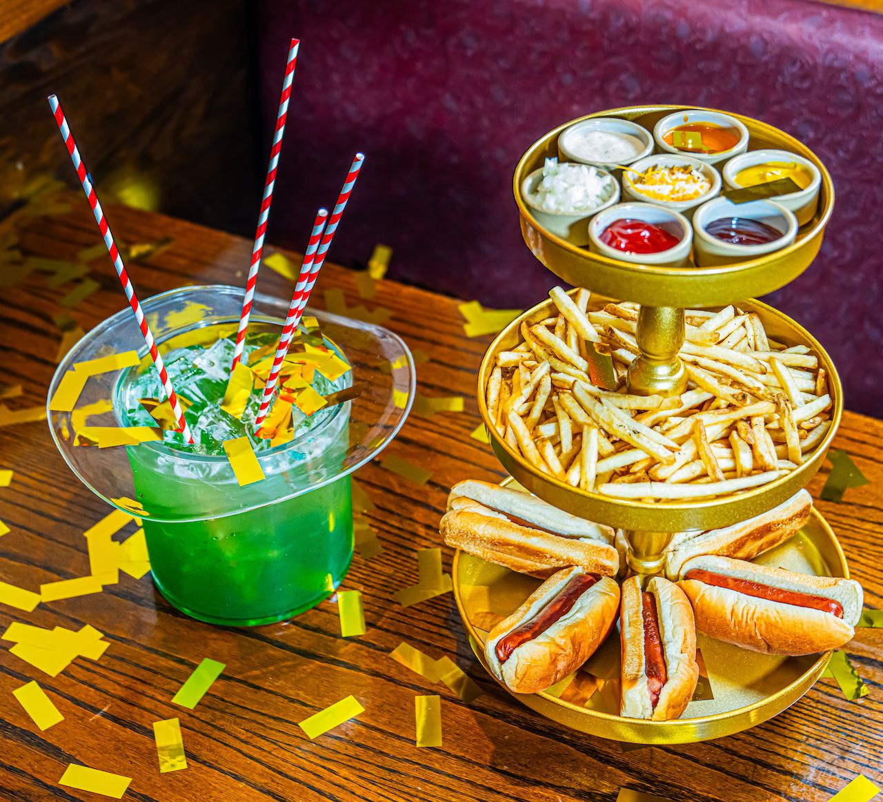 Bright green cocktail with red-and-white striped straws beside a gold tier tray of french fries and hot dogs; top holds small bowls of sauces, while confetti decorates a wooden table.