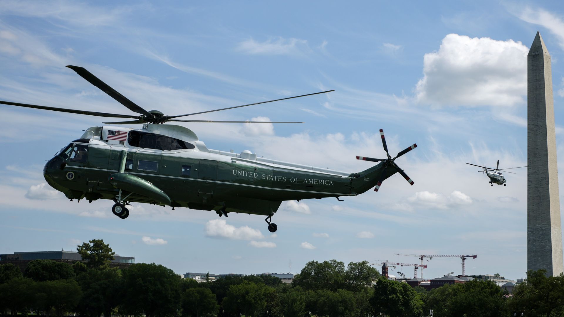 Green U.S. military helicopter labeled United States of America flies over a city with the Washington Monument in the background; a second helicopter is visible against a blue sky with wispy clouds.
