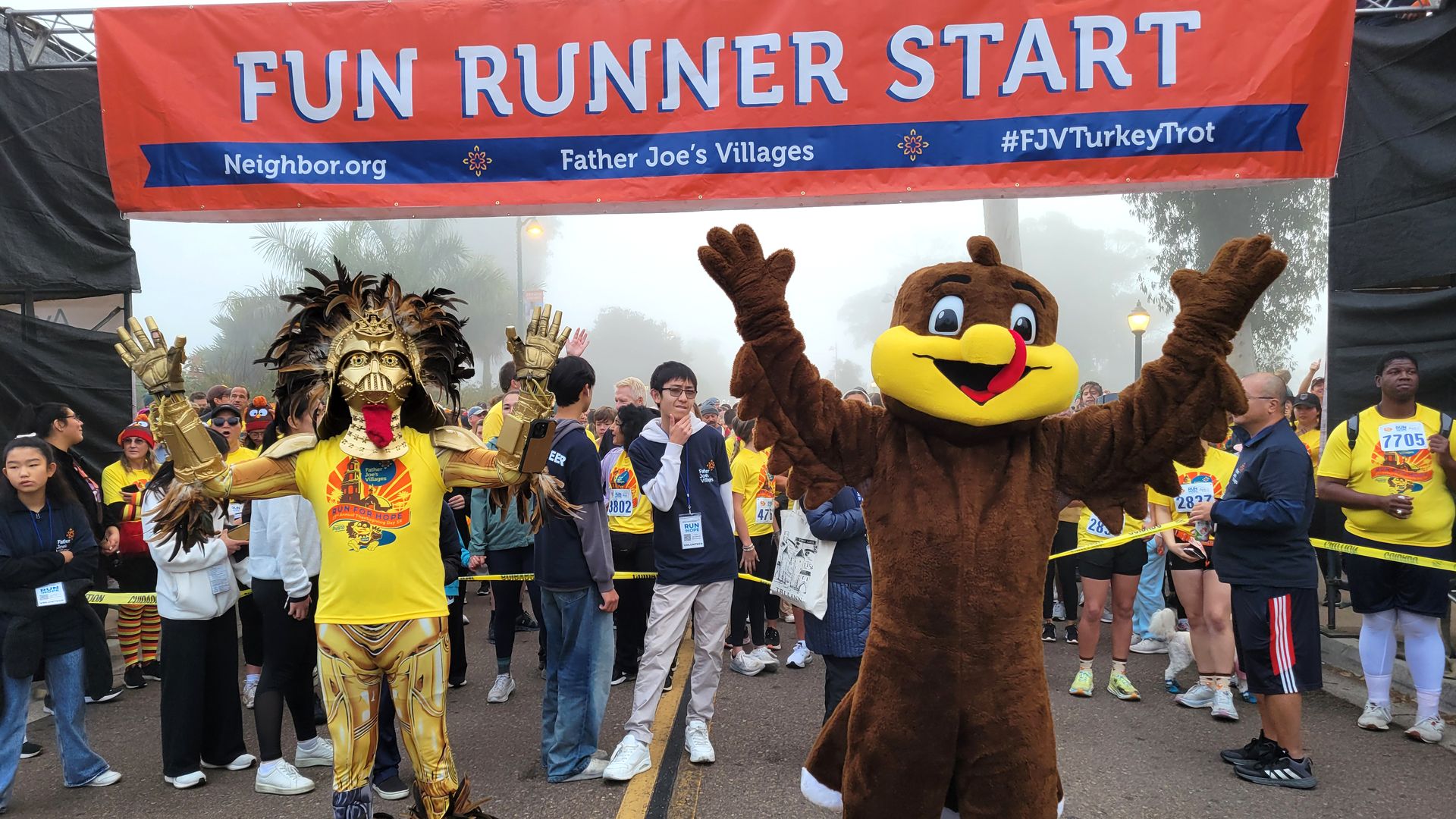 Crowd of runners under a red "FUN RUNNER START" banner, including a person in a detailed golden turkey costume and another in a large brown turkey mascot suit, raising their arms.