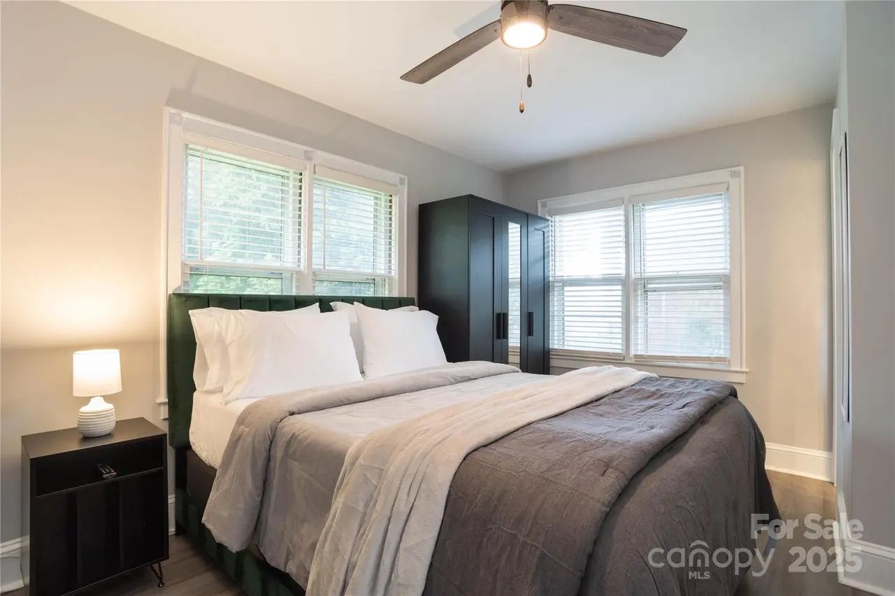 Bright bedroom with gray walls, large bed with white pillows and gray bedding, dark wood nightstand with lamp, black wardrobe, two windows with white blinds, and ceiling fan lighting.