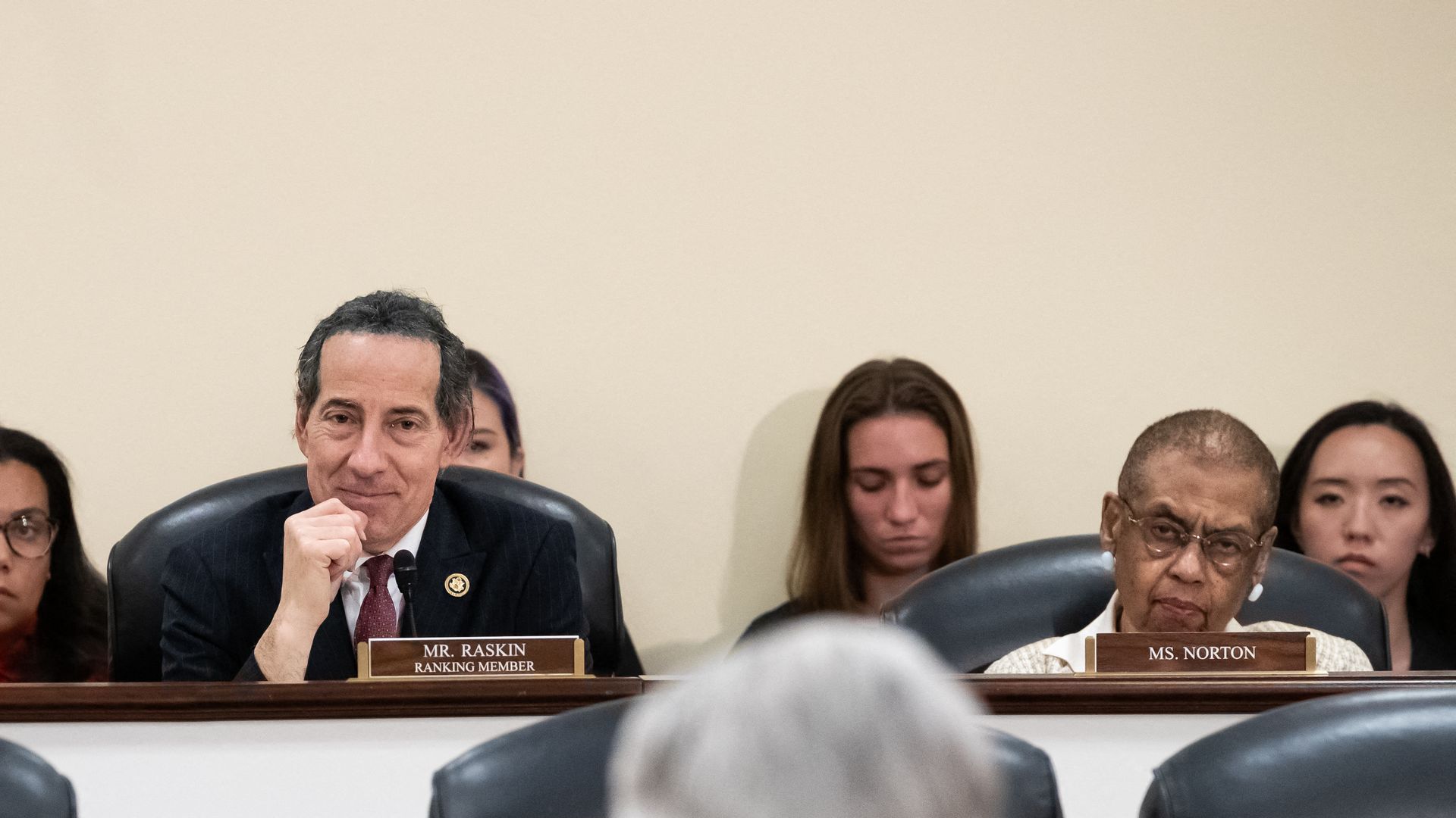 Panel of officials including Ms. Ocasio-Cortez, Mr. Raskin, and Ms. Norton seated behind a desk with nameplates in a formal setting, listening to a speaker facing them.
