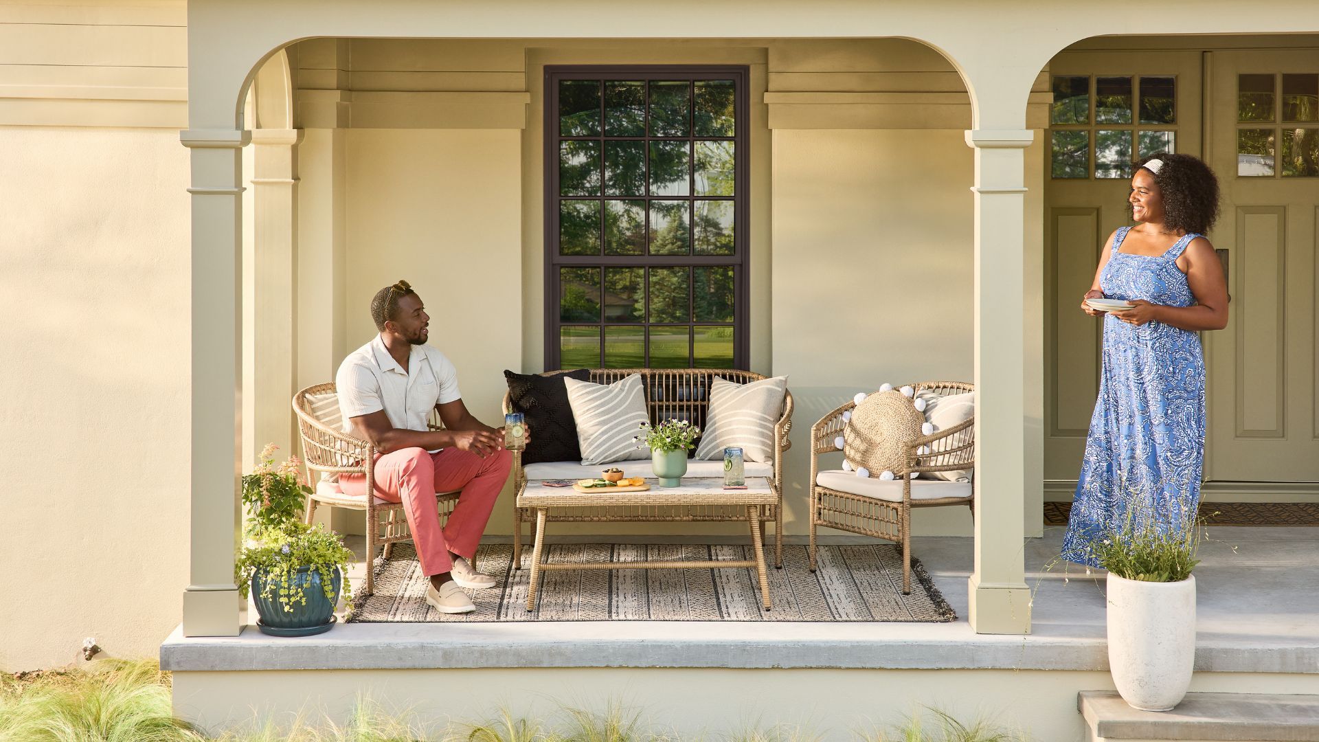 A man and woman enjoy a relaxed conversation on a front porch furnished with wicker seating and drinks on the table.