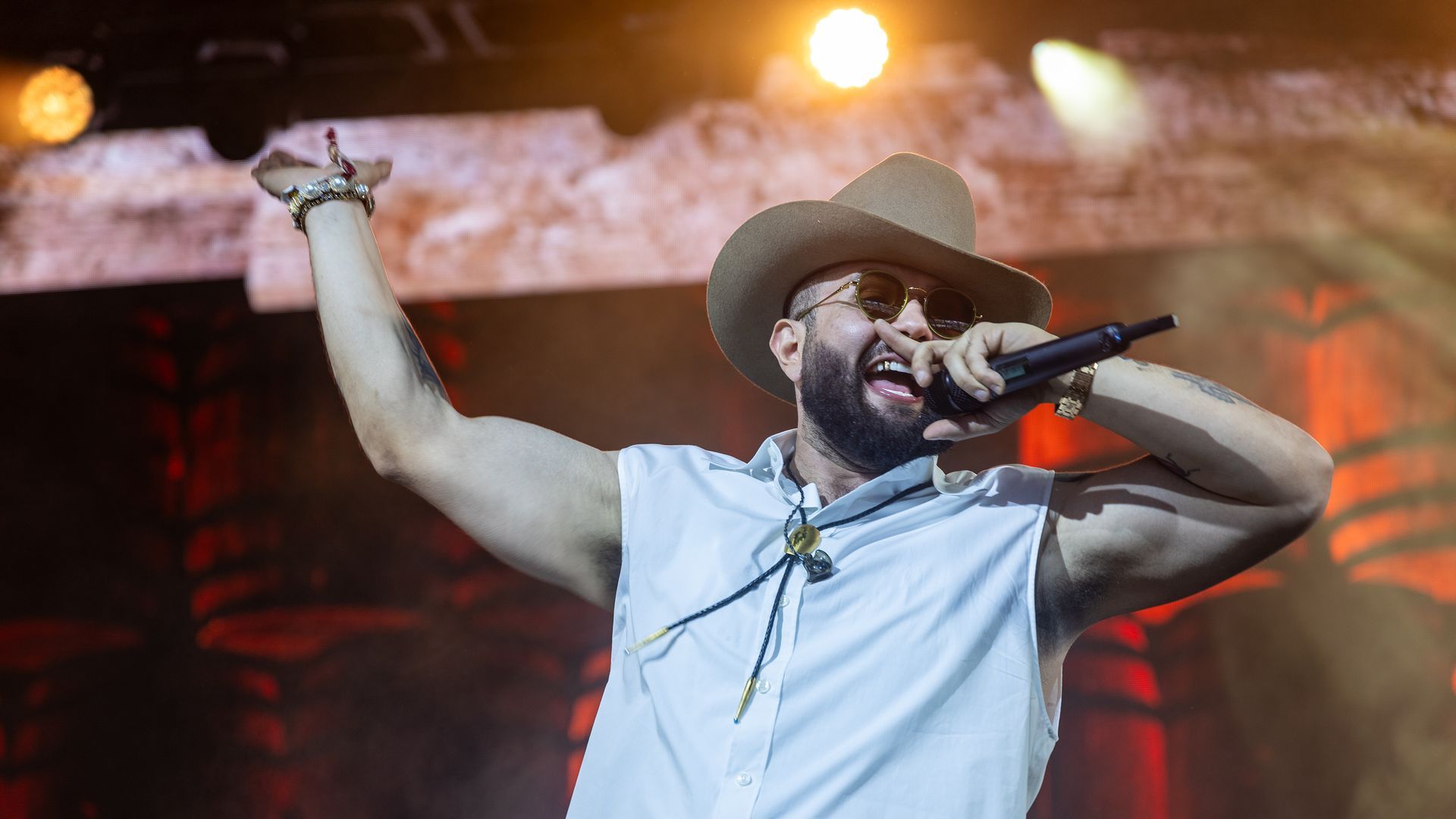 A man in a cowboy hat and sunglasses sings into a microphone.