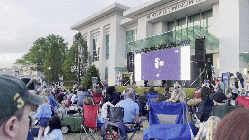 A crowd in front of Huntsville Museum of Art celebrates the splashdown of Artemis II mission by cheering and waving shakers. 