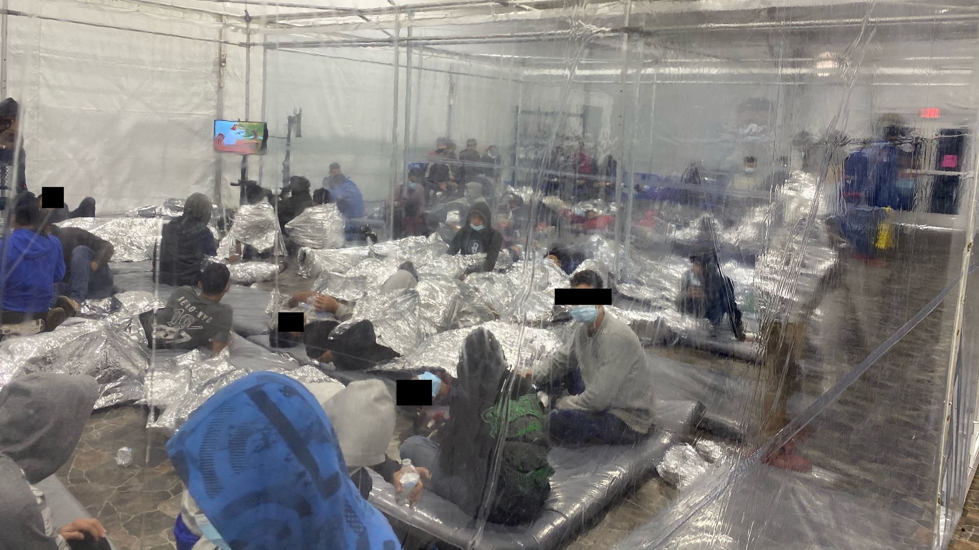 Migrants sit in a plastic contained area inside a tent, wrapped in aluminum blankets to keep warm