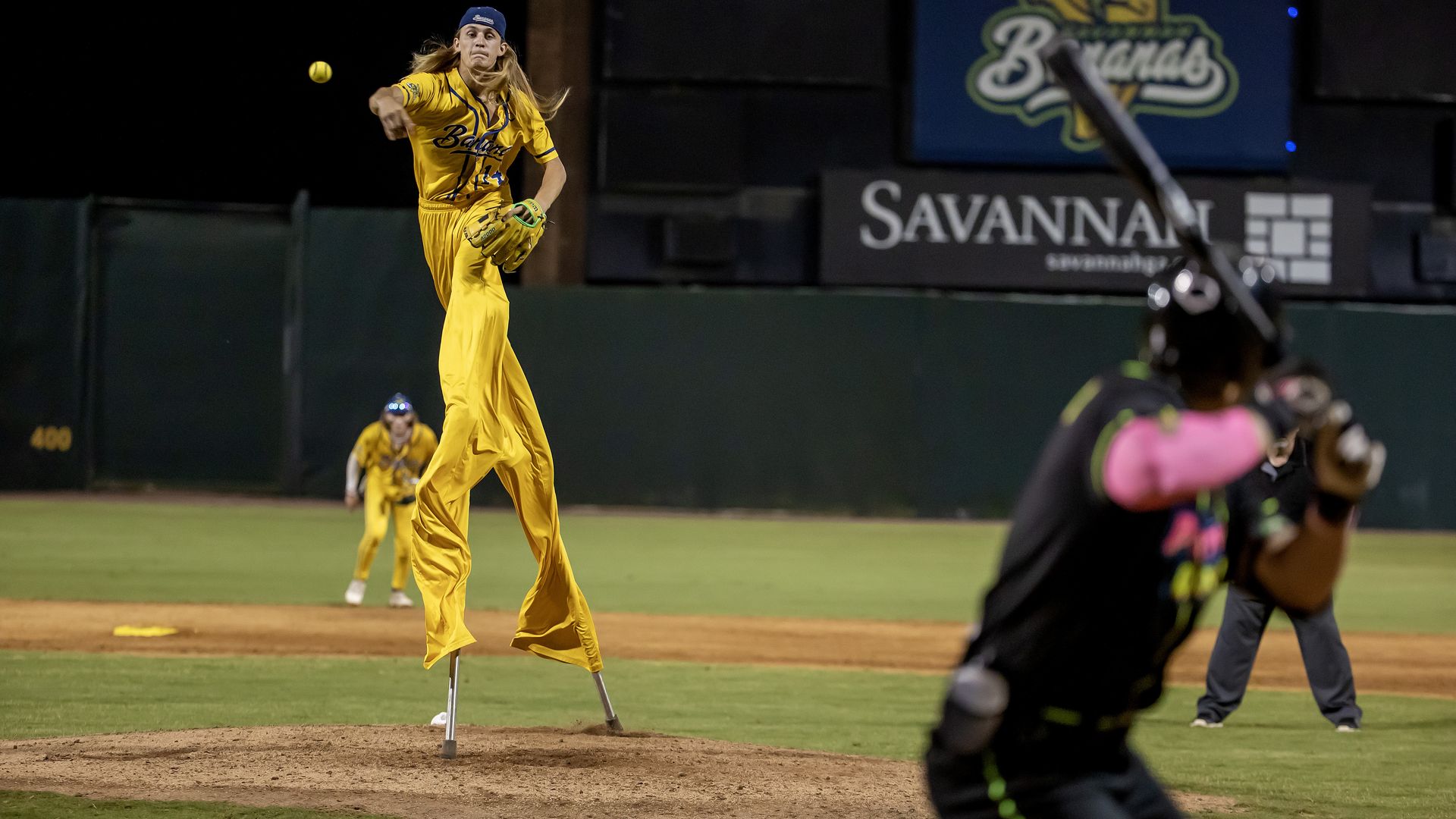 Night baseball scene: a female pitcher in a yellow uniform and blue cap throws from the mound; catcher and batter appear in the foreground, with a Savannah sign on the outfield wall.