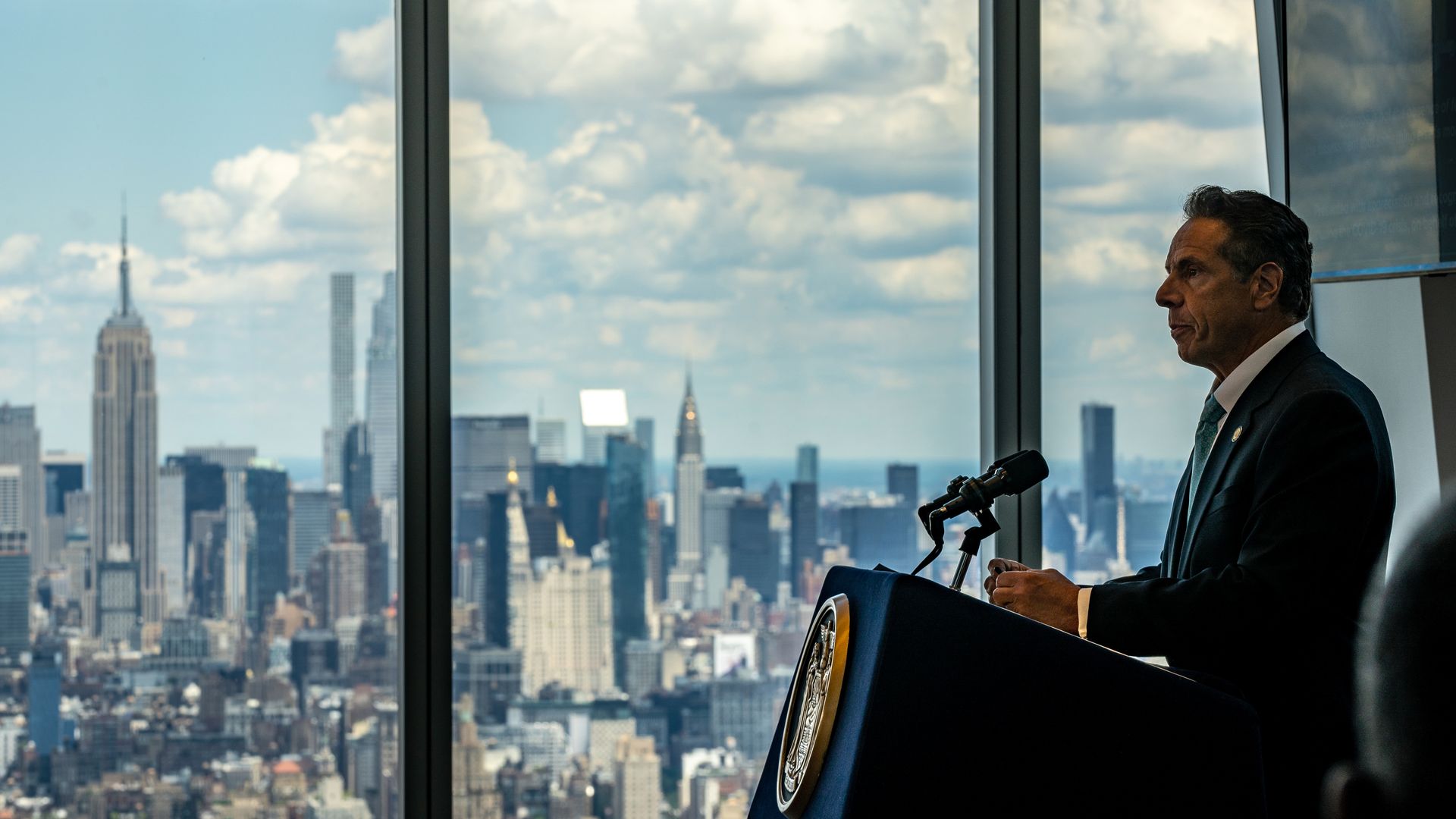 Andrew Cuomo stands at a lectern with a large window behind him showing the Manhattan skyline 