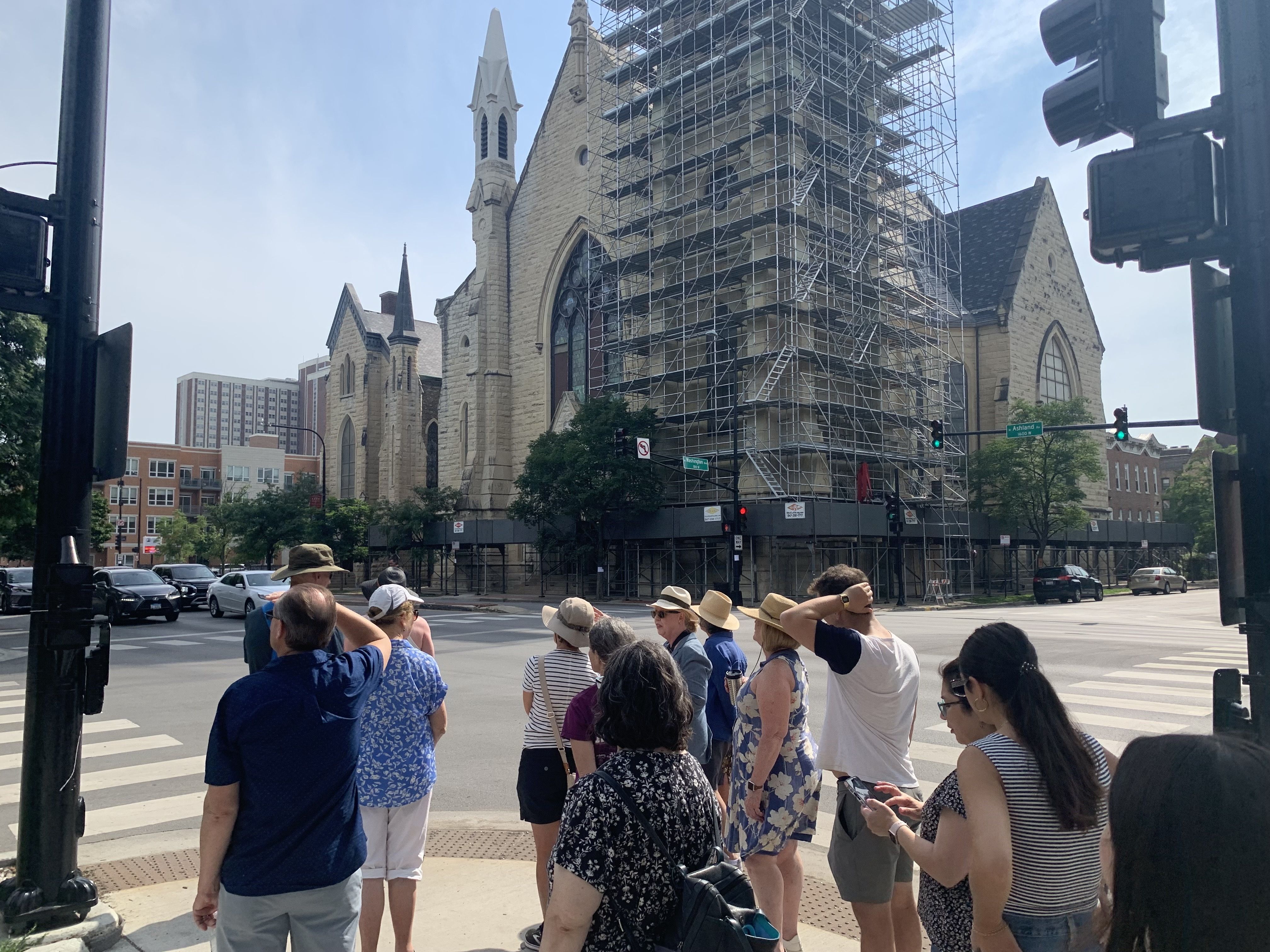 A group of people waiting at a crosswalk near a stone church under construction with scaffolding on a sunny day in an urban area.