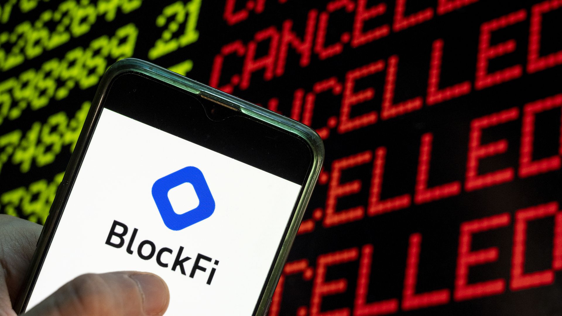 In photo-illustration, phone with BlockFi logo held in front of a sign that says Cancelled