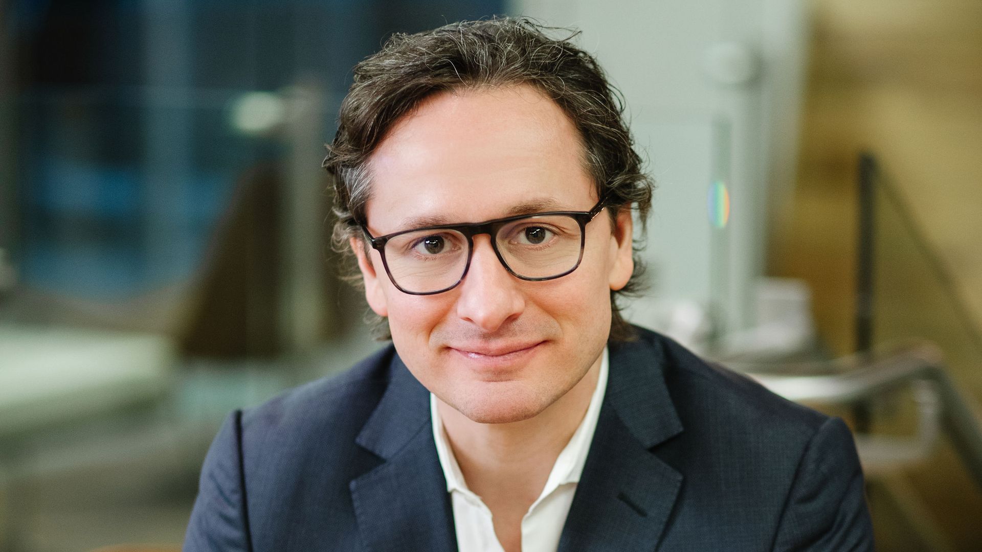 Portrait of a Jim O'Leary in a navy suit and white shirt, wearing black glasses, seated with hands folded. Slight smile in a modern office setting with a blurred background.