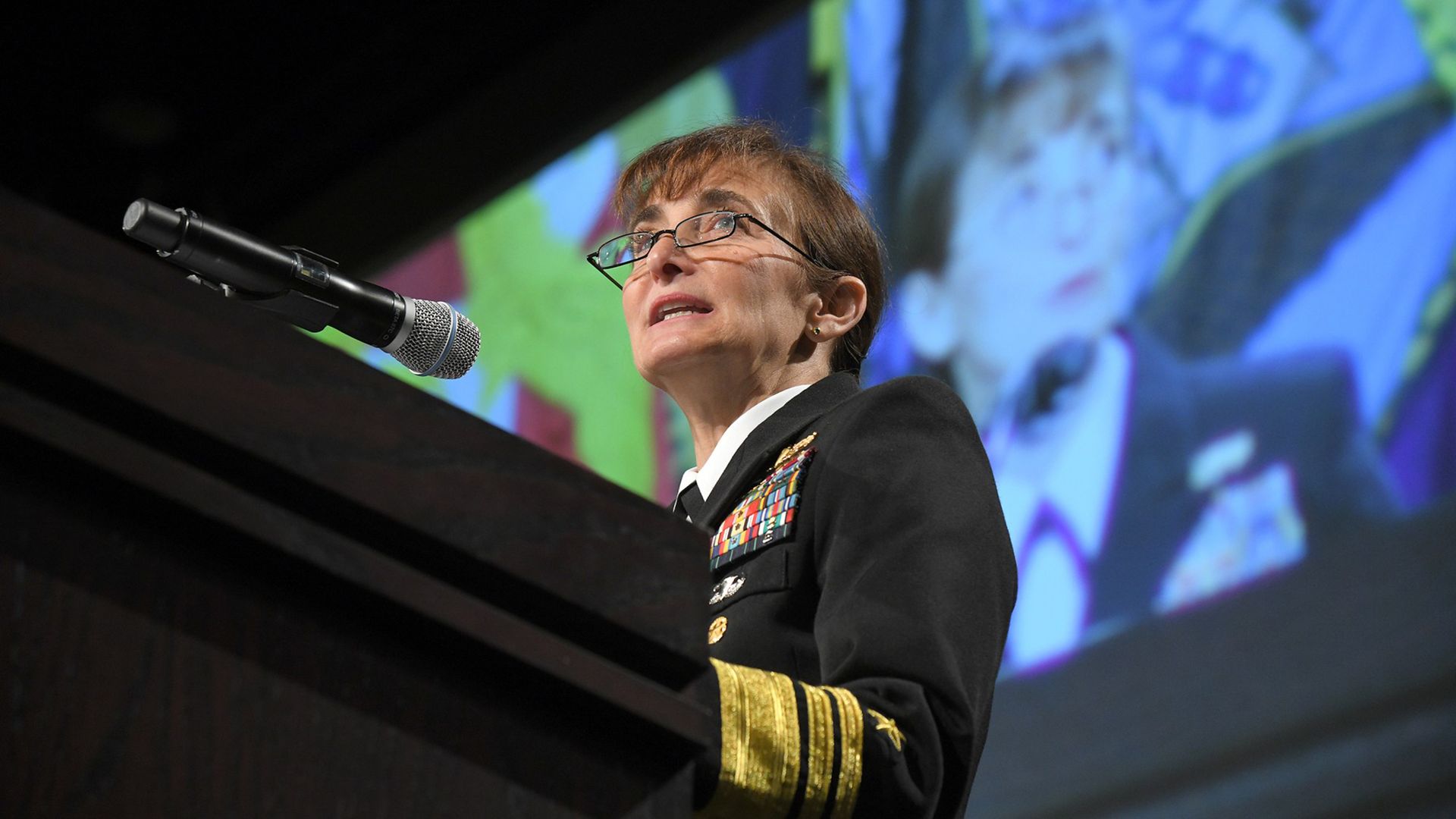Female military officer in black uniform with gold stripes and medals speaks into a podium microphone, with a large colorful projected image behind her.
