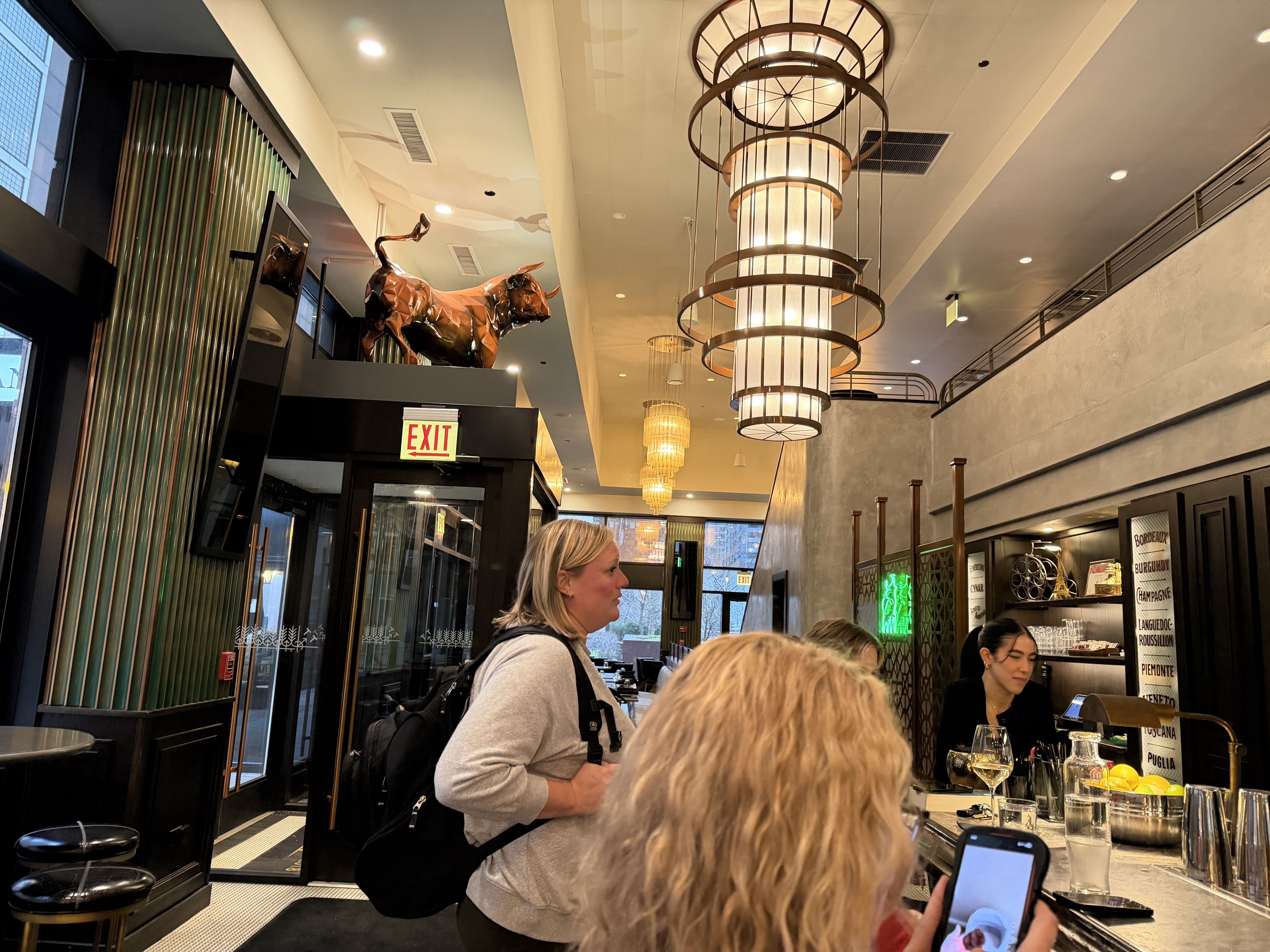 Chic restaurant bar with a metallic horse sculpture above a doorway, green striped walls, and large round pendant lamps. A blonde woman with a backpack stands at the bar.