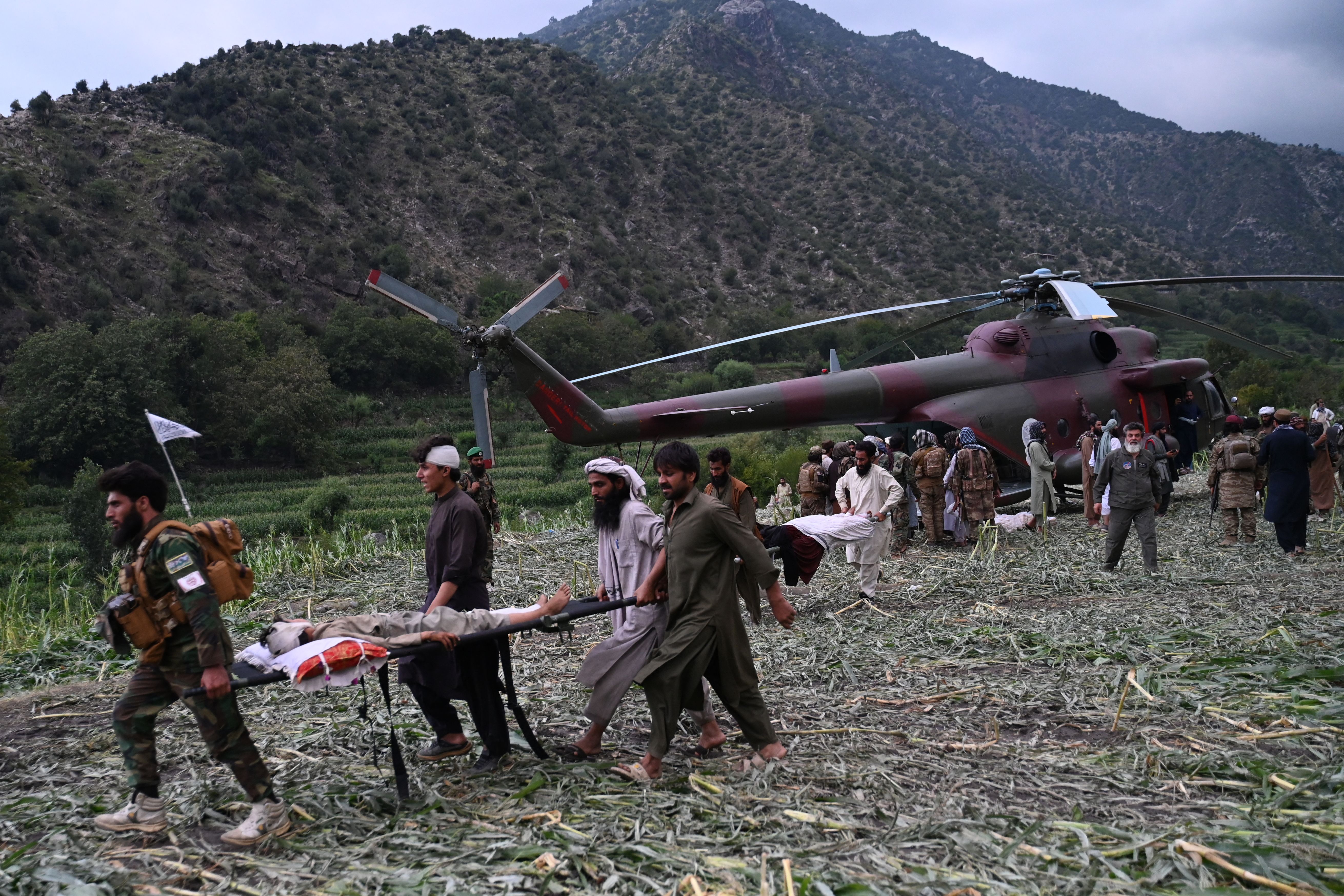 Men in traditional Afghan dress carry people on stretchers near a military helicopter beside a hill.