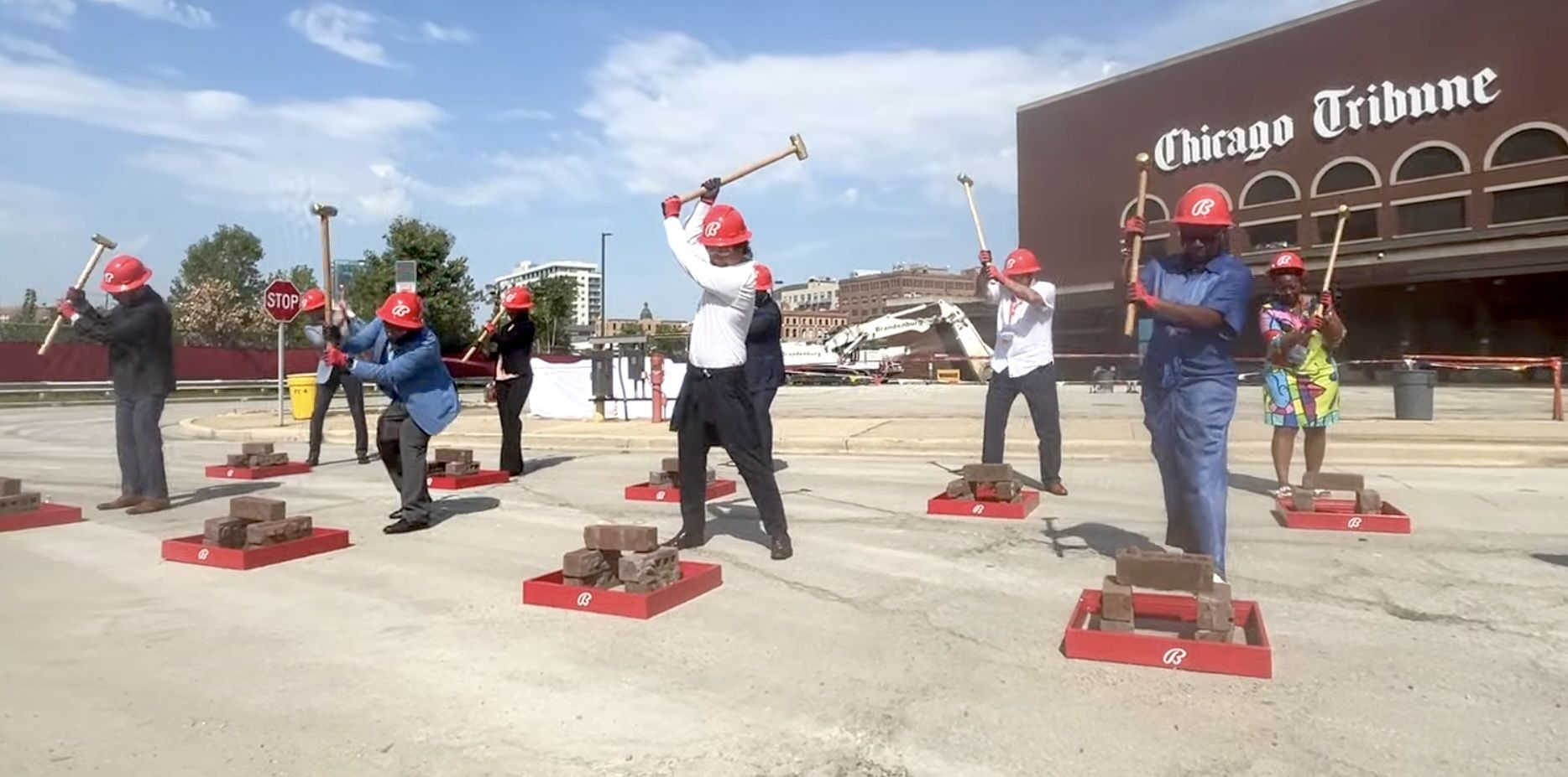 People in red hard hats in front of bricks raising sledgehammers to bricks.