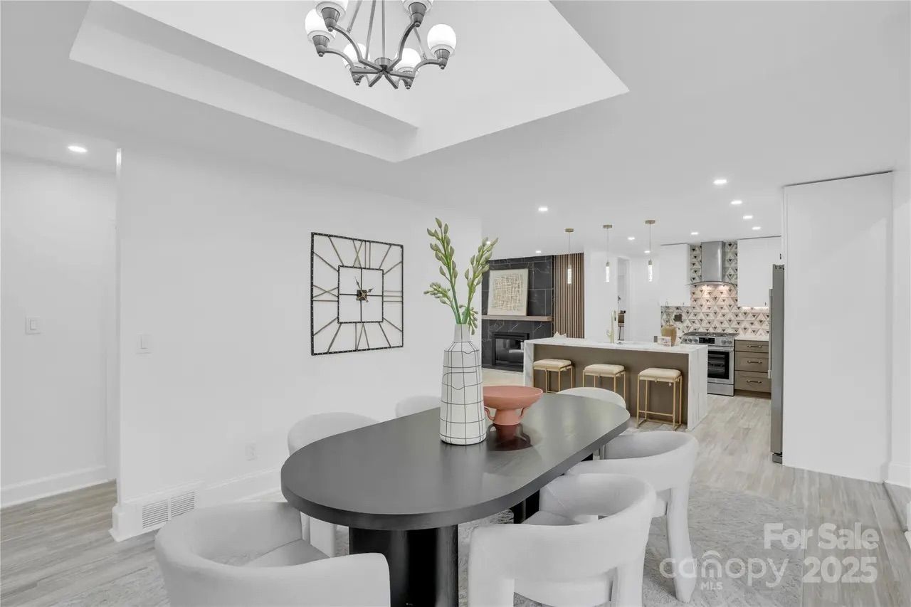 Modern dining area with black oval table, six white chairs, decorative vase, chandelier, and open kitchen featuring island with stools, stainless steel appliances, and patterned tile backsplash.