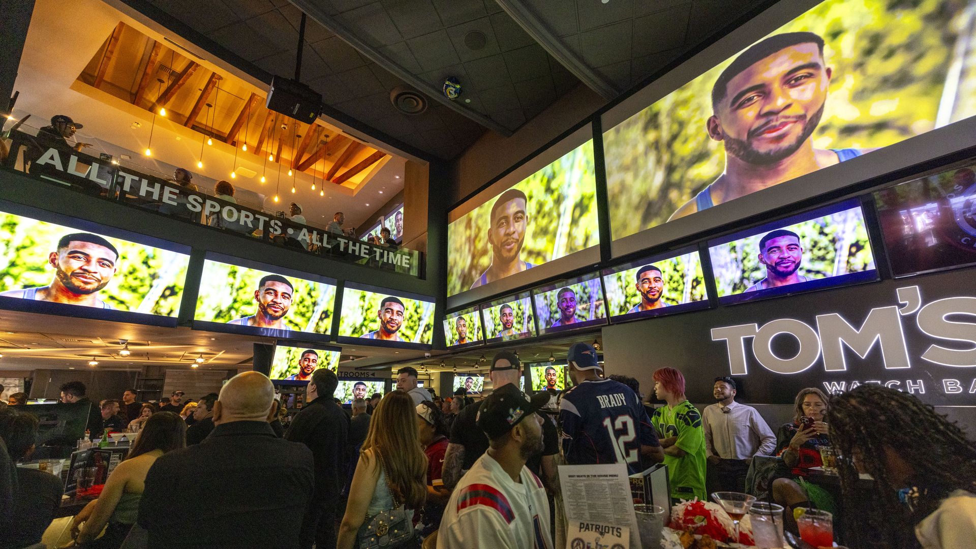 Crowded sports bar with multiple screens showing a person in a blue tank top outdoors. People wear sports jerseys, some seated with food and drinks, beneath a sign reading "TOM'S".