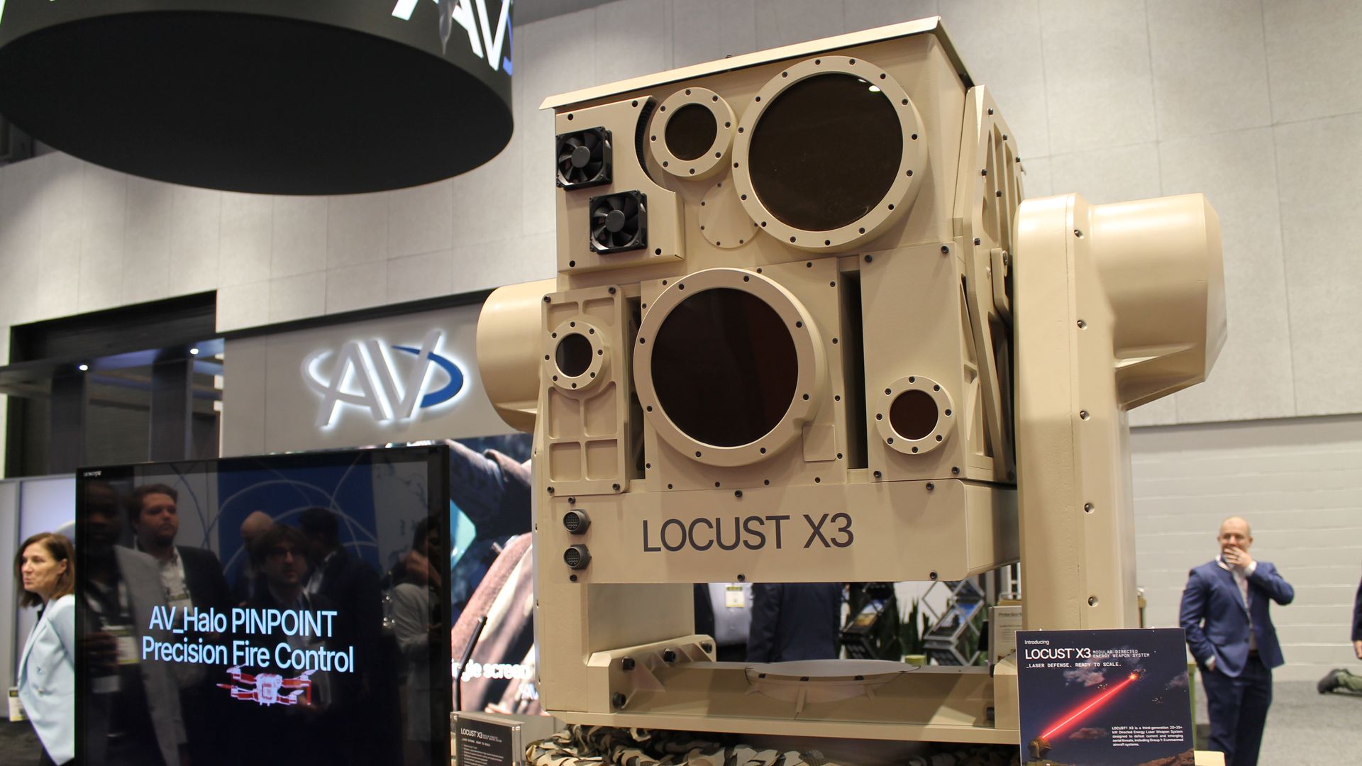 Beige LOCUST X3 military platform with large circular ports, fans, and tracked base on display at a convention. A screen shows "AV Halo PINPOINT Precision Fire Control" as attendees observe.
