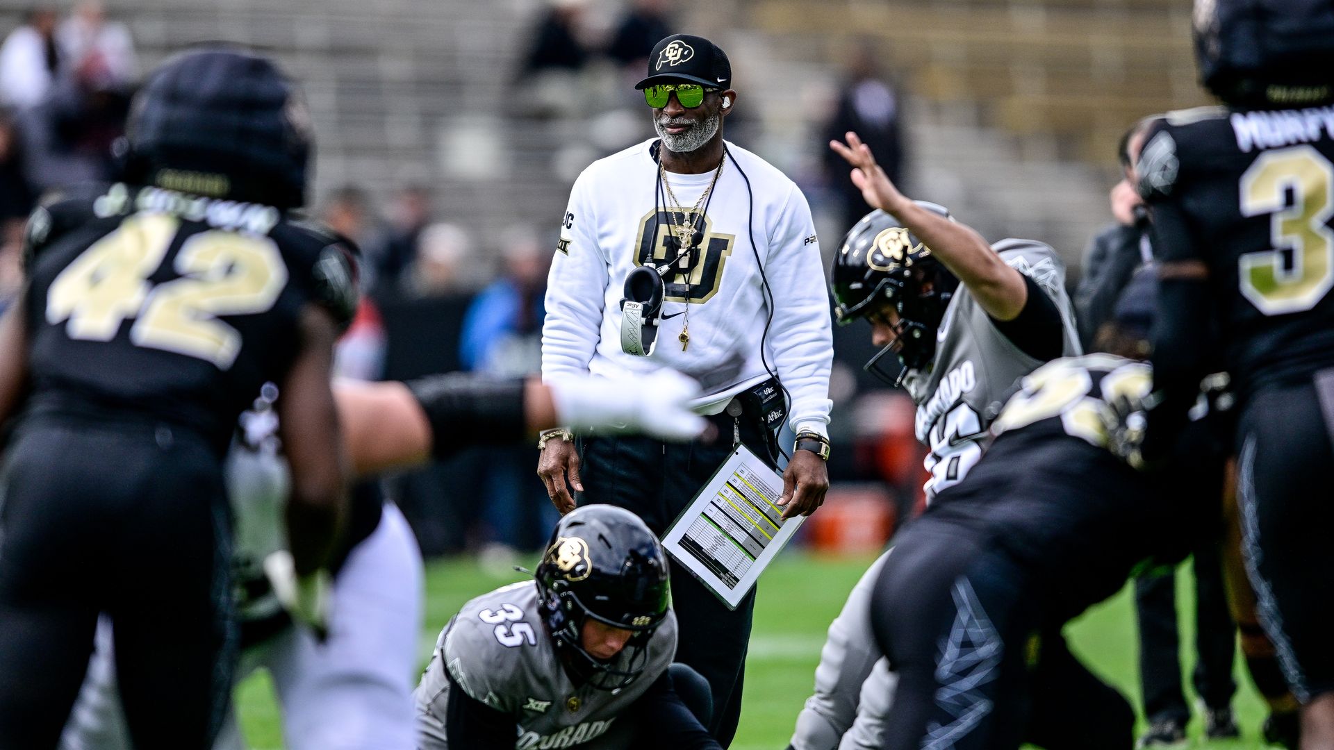 Football coach in white CU sweatshirt and black cap watches players in black and gray Colorado uniforms during a field goal attempt on a green field.