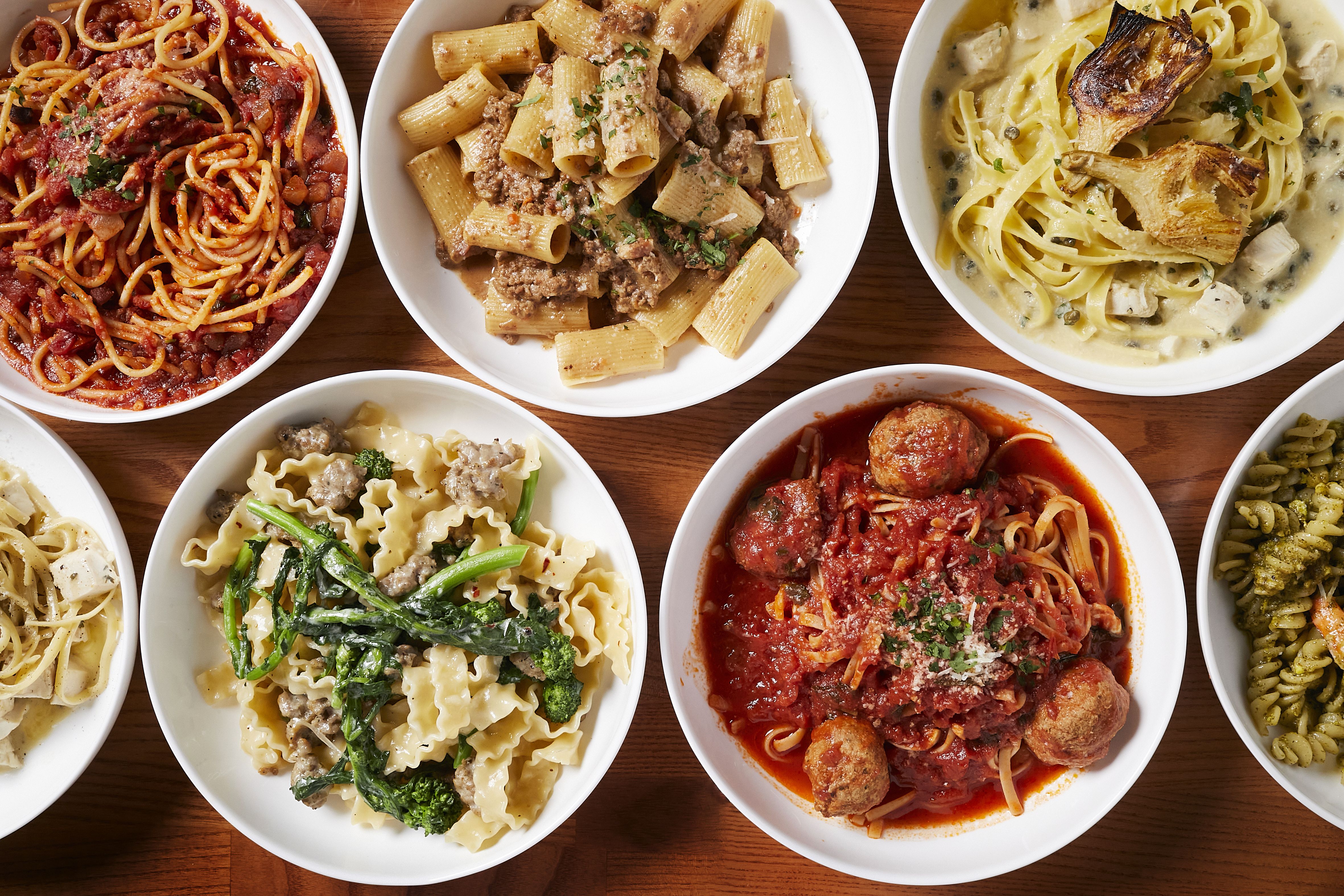 Top-down view of many white bowls of pasta on a wooden table: spaghetti with tomato sauce on the left, rigatoni with meat sauce center, and fettuccine alfredo with mushrooms on the right.