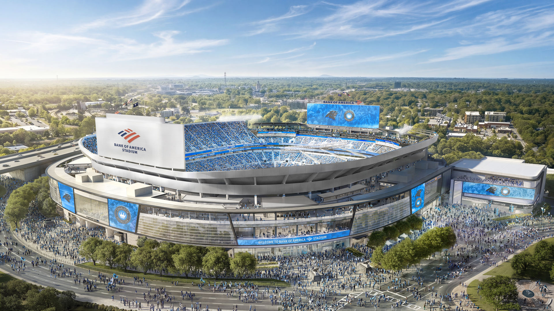 Panoramic view of Bank of America Stadium with blue seating, a large digital scoreboard, and crowds outside. Bright day, surrounding green trees and distant cityscape.