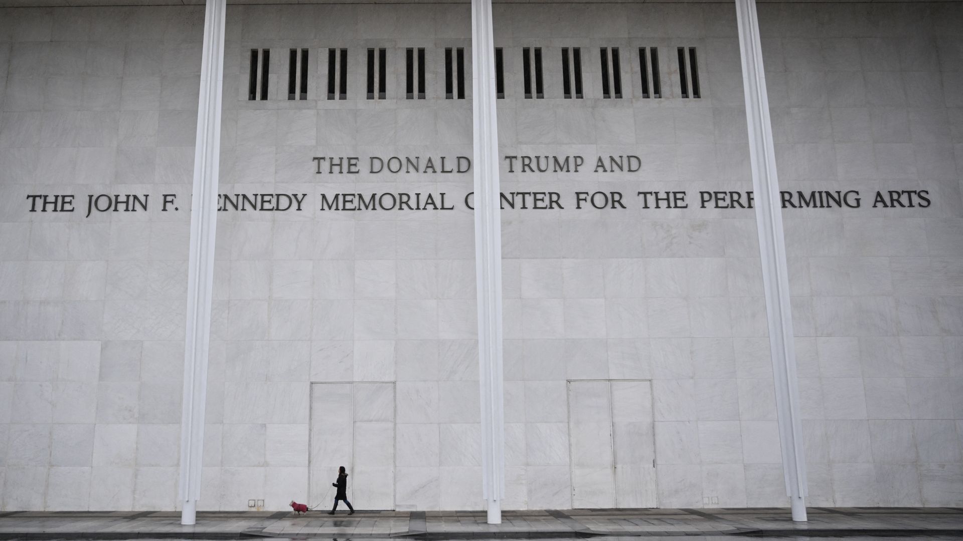 Woman walking a small dog in front of a large white marble wall with the inscription "The Donald Trump and The John F. Kennedy Memorial Center for the Performing Arts."