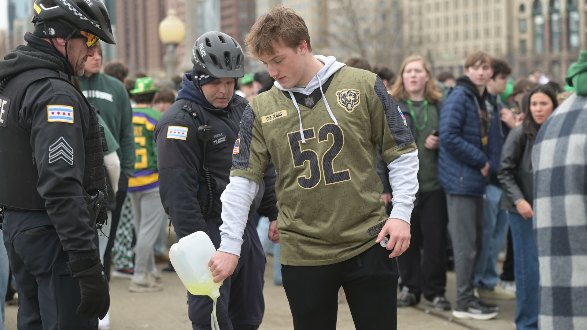 Young man wearing an olive-green Chicago Bears jersey with the number 52 pours a pale yellow liquid from a jug as police officers stand nearby and a crowd watches on a city street.