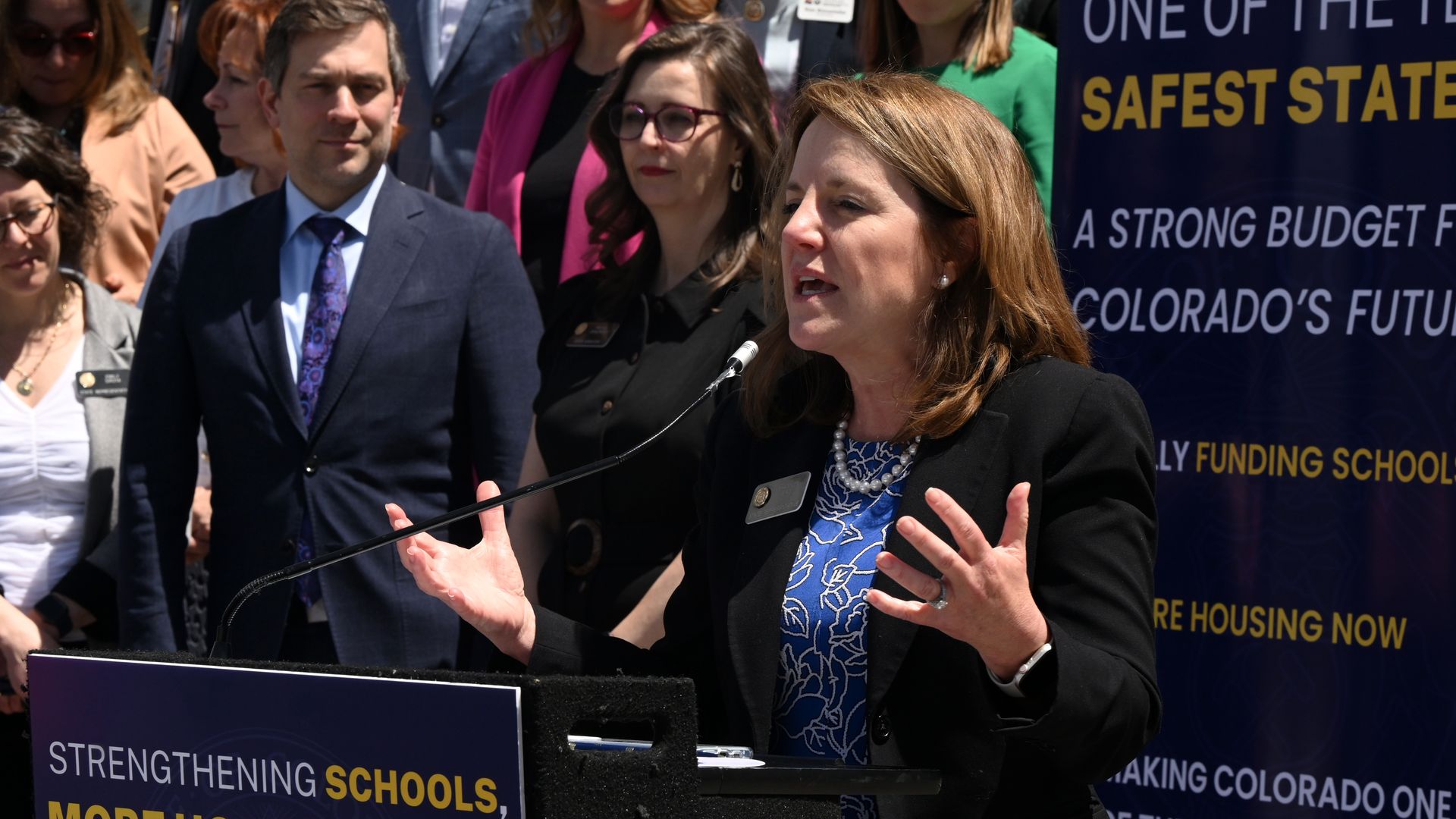 A woman extends her hands while speaking at a lectern. 