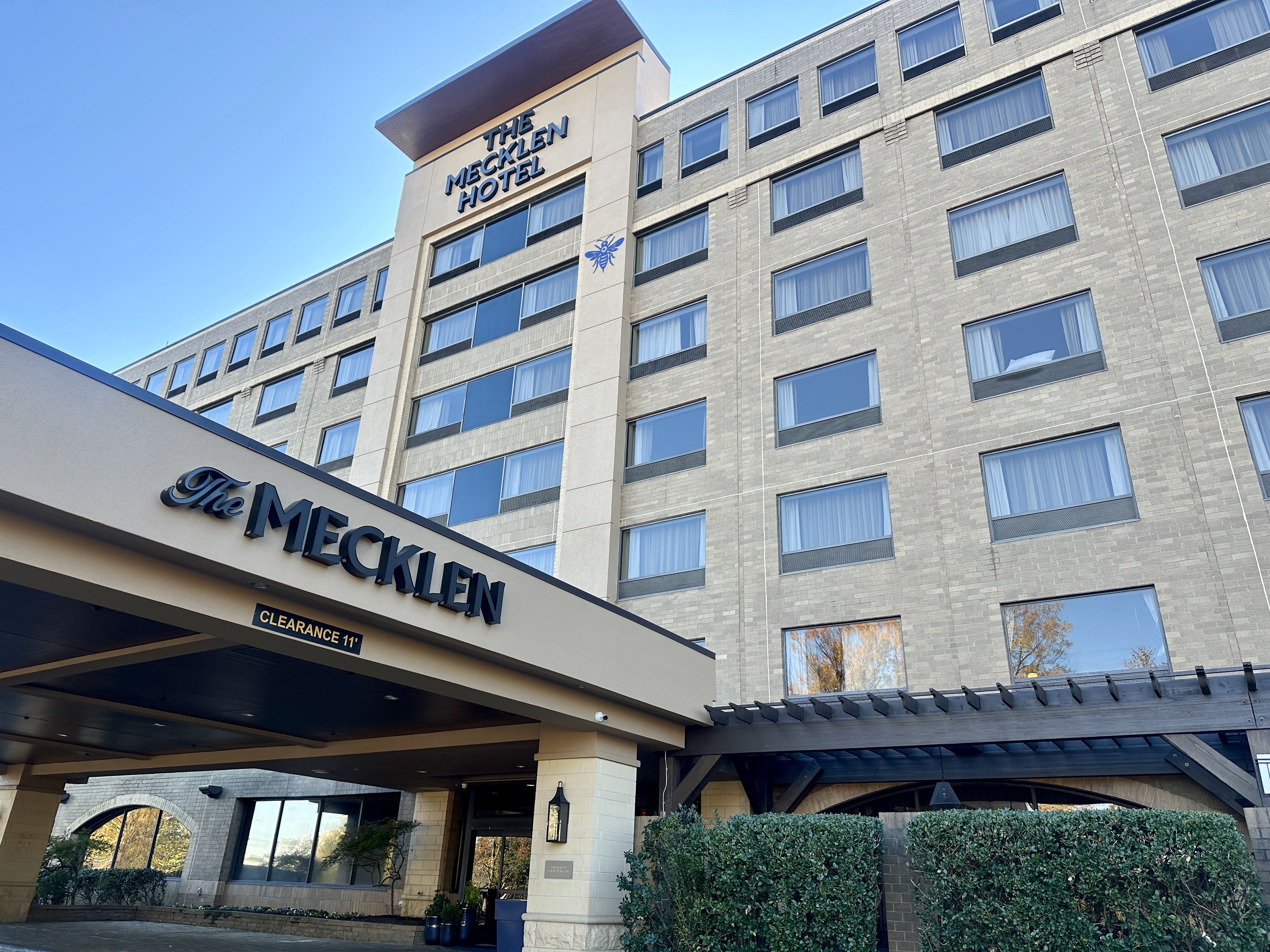 Exterior view of The Mecklen Hotel, a multi-story beige brick building with many windows, a covered entrance with the hotel name, clear blue sky, and some green bushes in front.