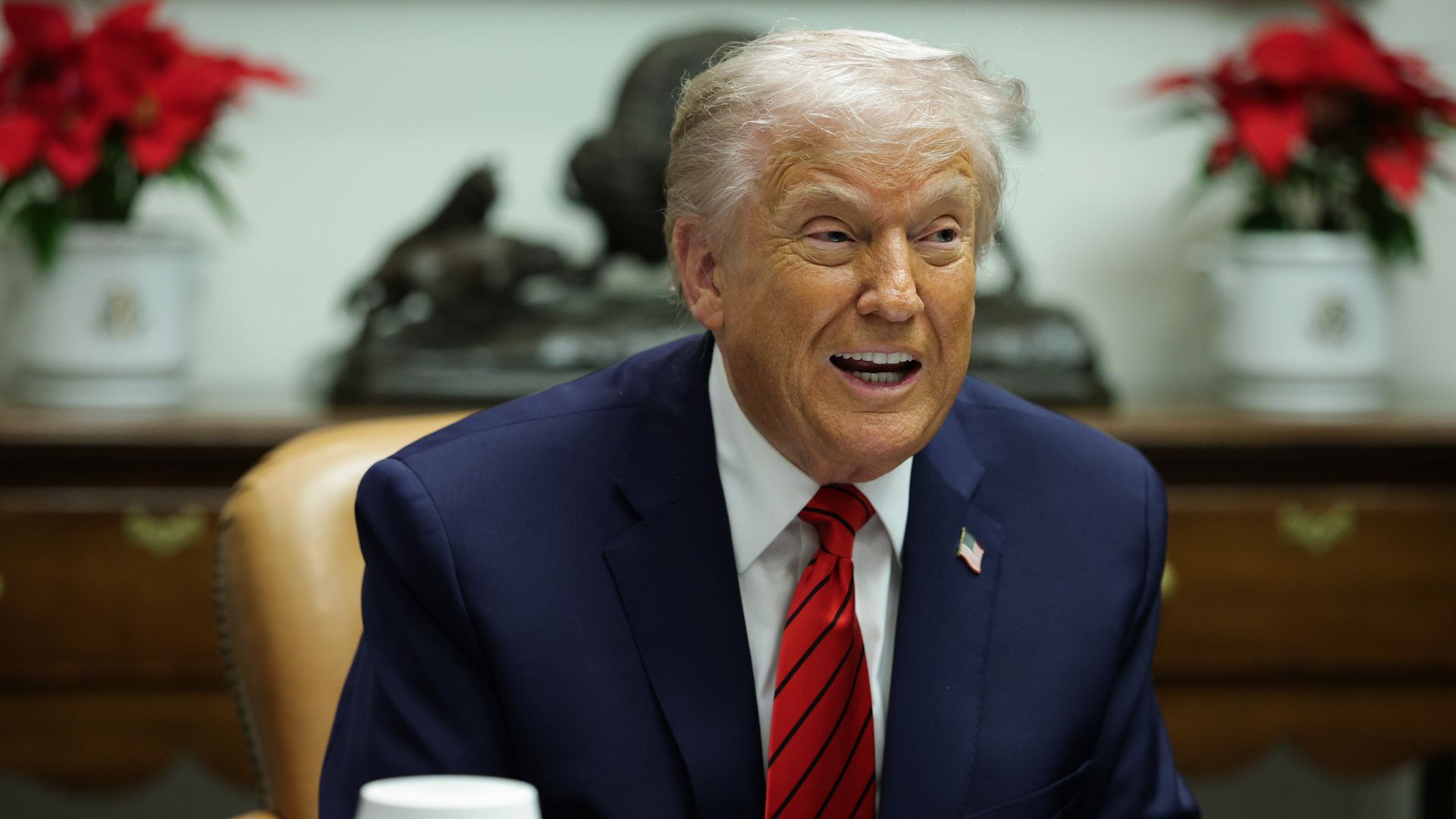 President Trump — wearing a dark blue suit, a red tie, a white collared shirt and an American flag pin — speaks during a White House roundtable discussion while sitting in a light brown chair.