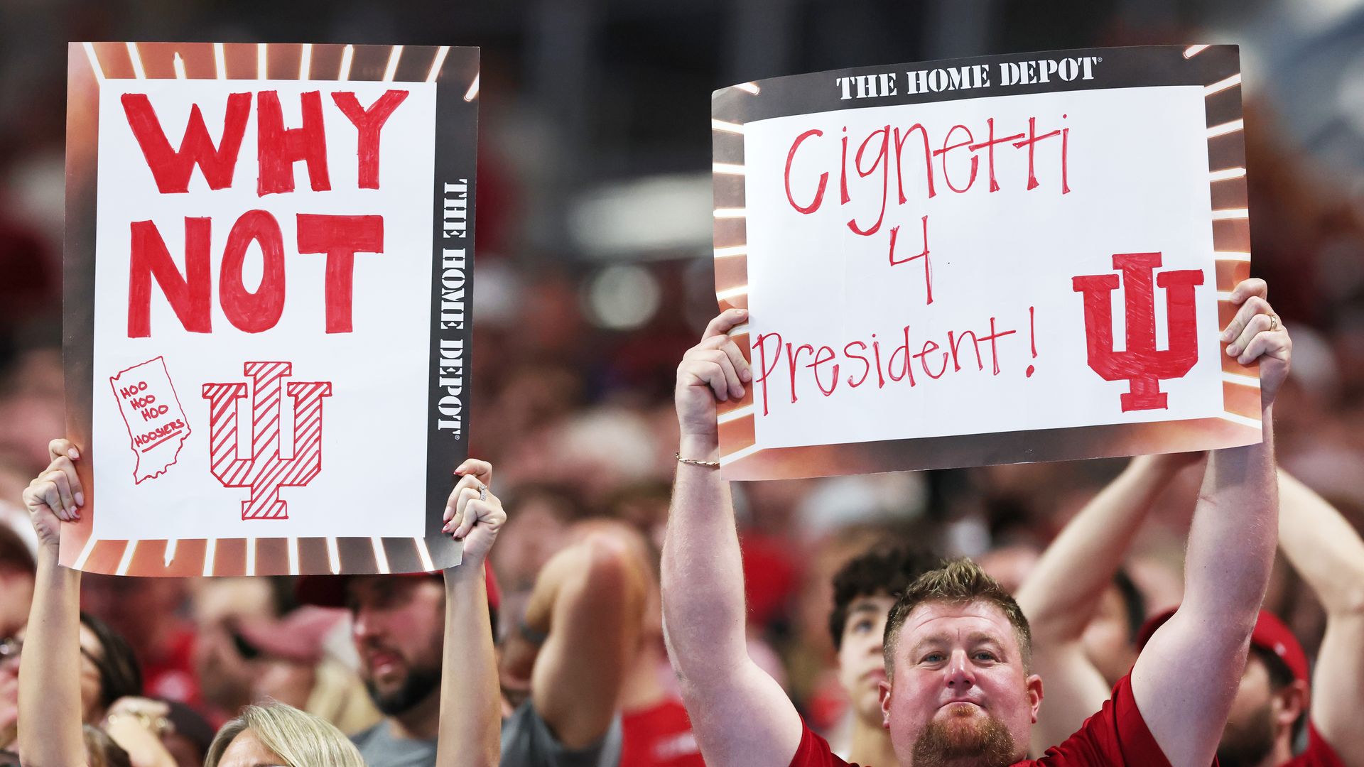 Two Indiana University fans in red hold signs: one says "WHY NOT" with IU logo, the other says "Cignetti 4 President!" with IU logo, at a crowded sports event.