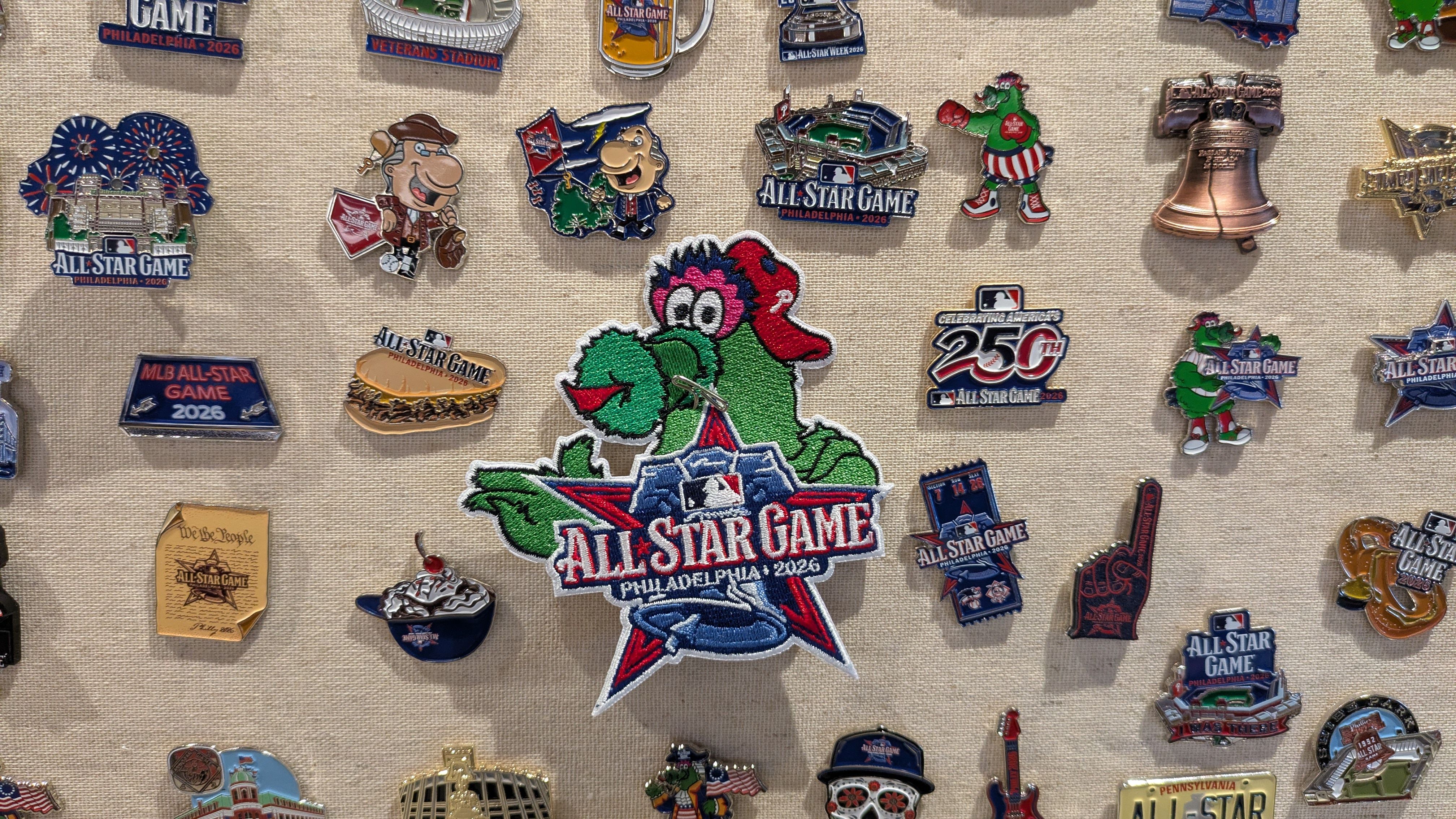 Display of MLB "All-Star Game" pins on a beige fabric board, featuring Philadelphia 2026 logos, mascots (including the Phillie Phanatic), trophies, stars, and various All-Star badges and patches.