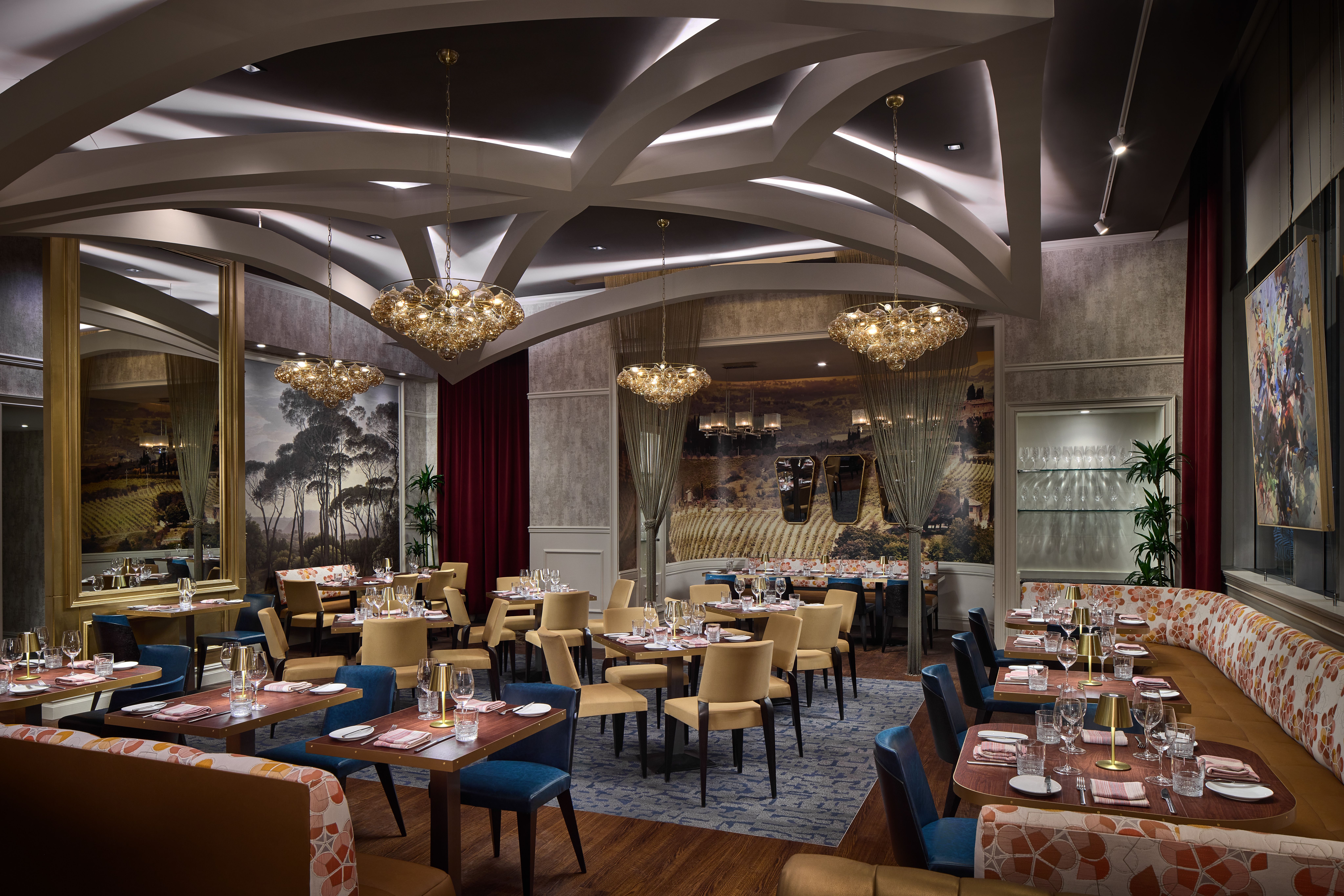 Elegant restaurant dining room with white arched ceilings and gold chandeliers. Tables set with glassware and pink napkins; beige and blue chairs, a long floral sofa, vineyard murals, and plants.